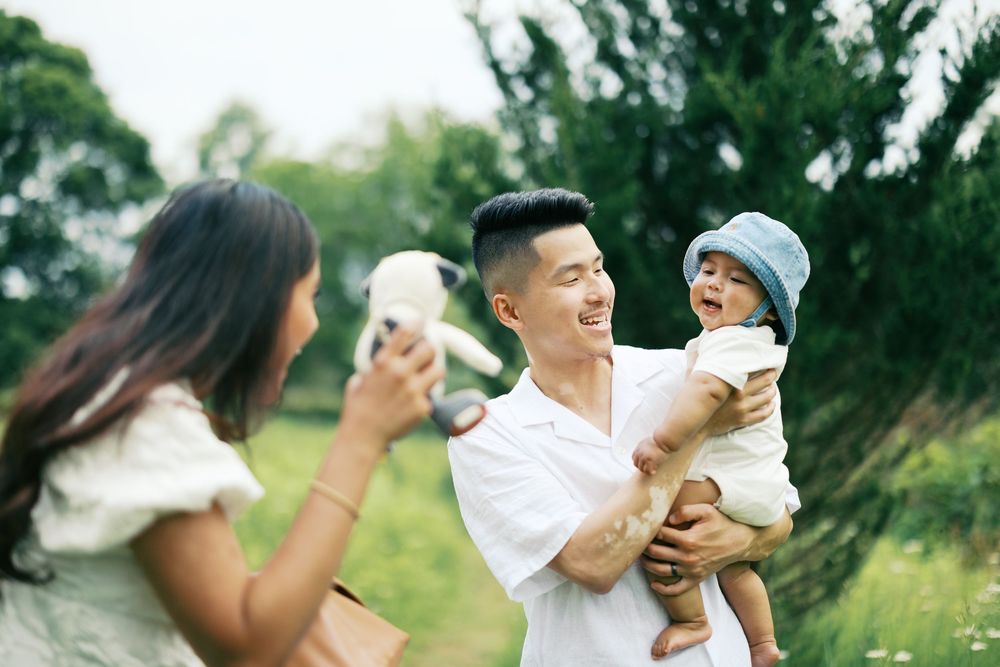 Happy parents in casual clothes enjoying drinks at the park with their baby wearing a blue hat on a sunny day.