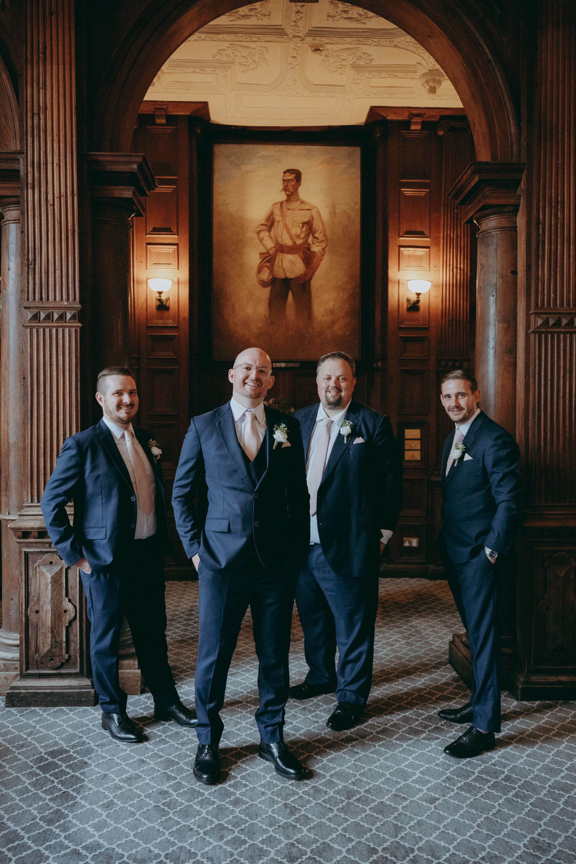 A sequence of five groomsmen in dark suits posing playfully in a grand hallway with ornate columns and lighting.