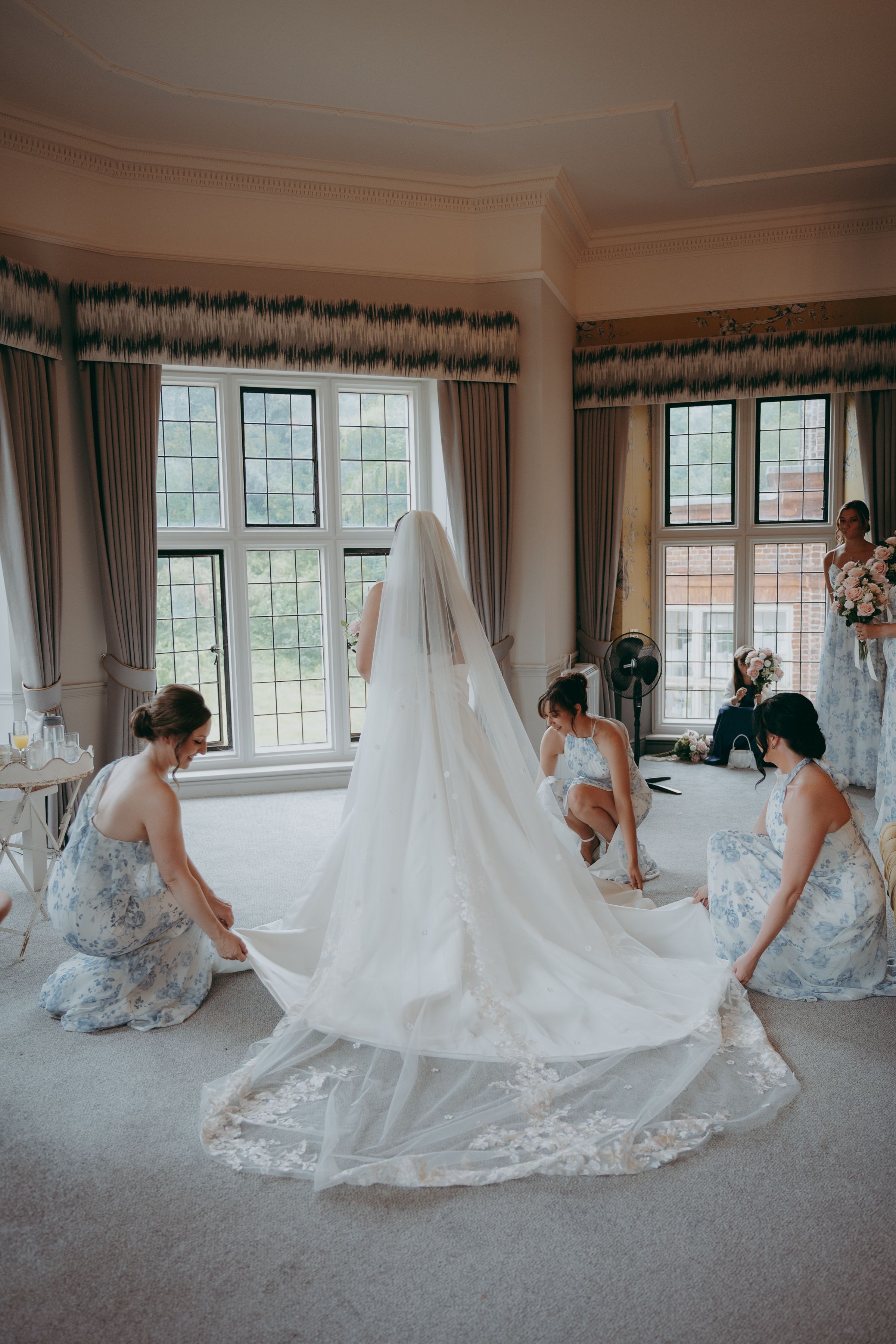 A wedding party helps adjust a long flowing cathedral veil near bright windows in an elegant room with green valances.