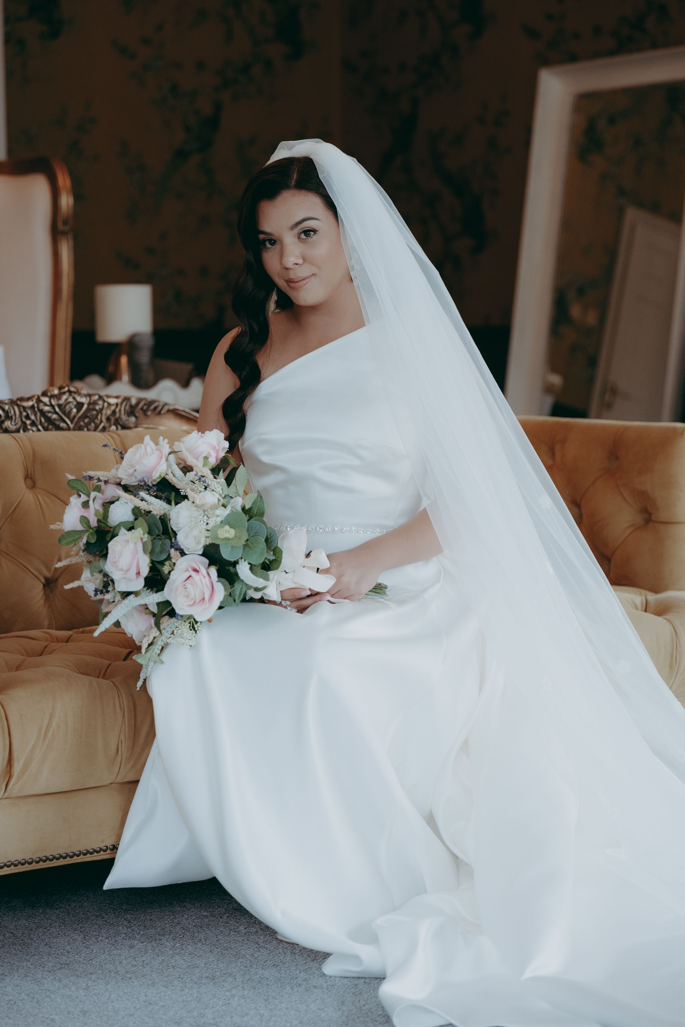 A bride in a white wedding gown poses elegantly with a bouquet of pink and white flowers on a beige sofa.