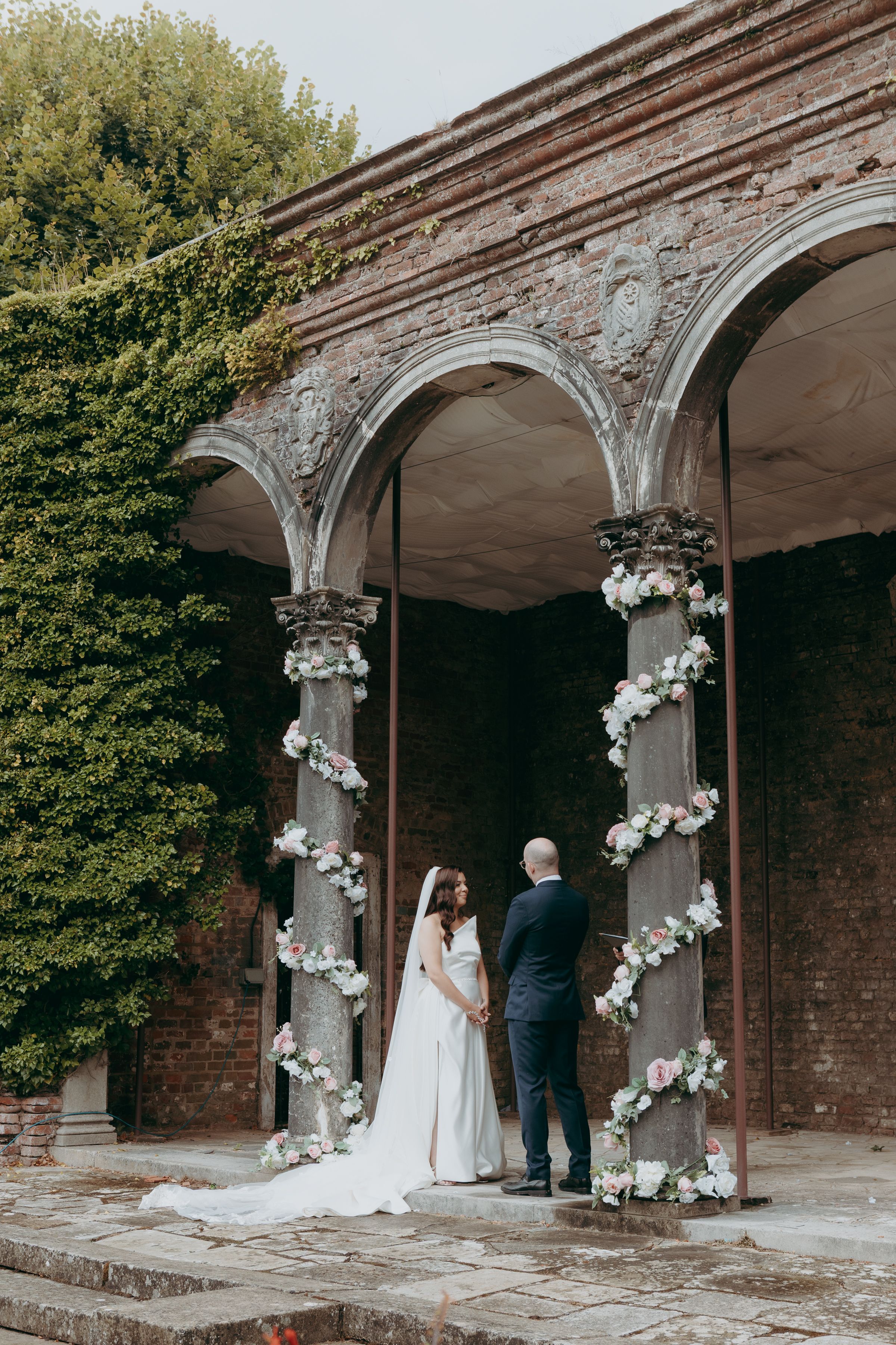 A historic brick archway decorated with white floral arrangements creates a romantic wedding ceremony backdrop.