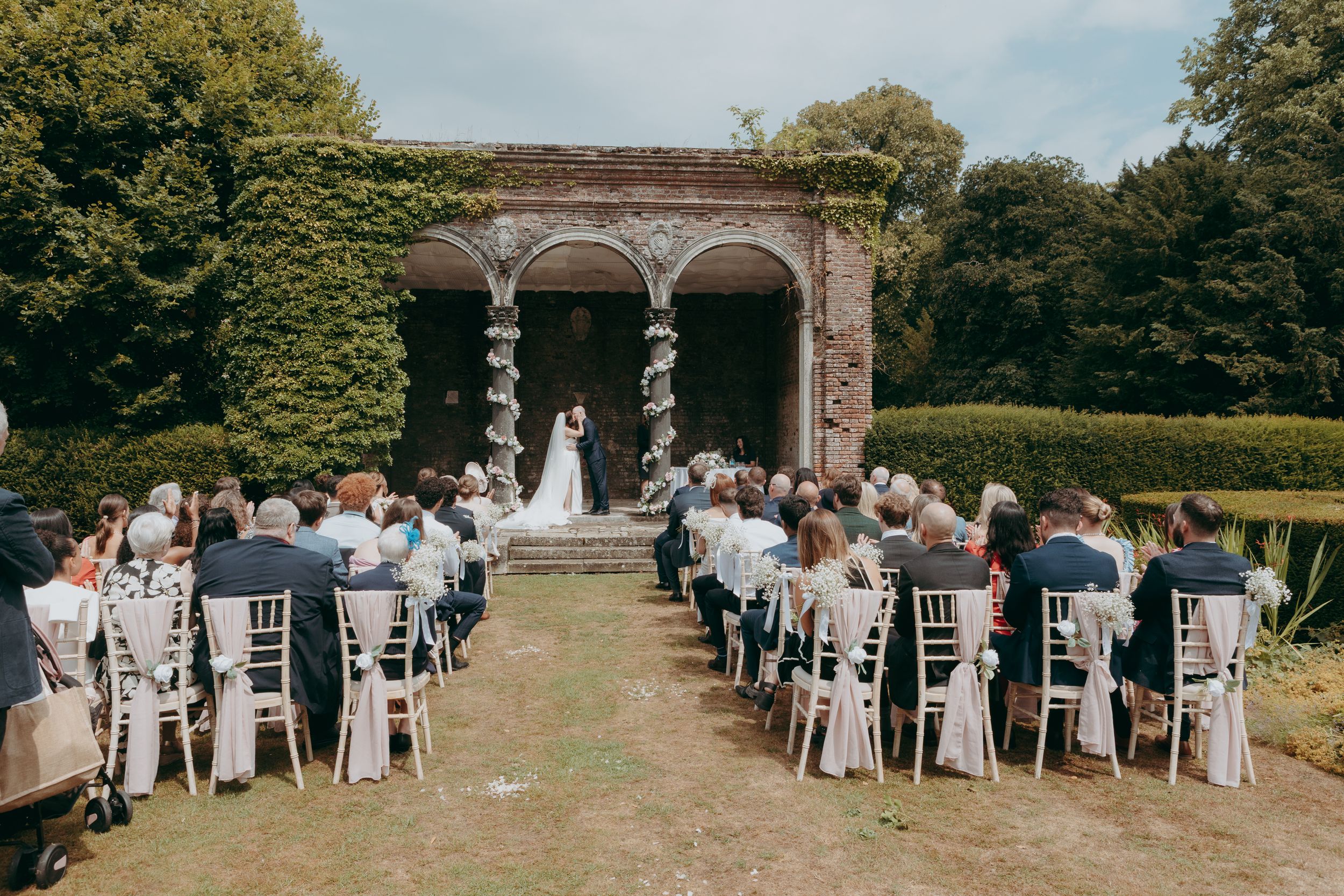 Outdoor wedding ceremony setup with white chairs arranged in rows along a tree-lined path leading to a brick archway.