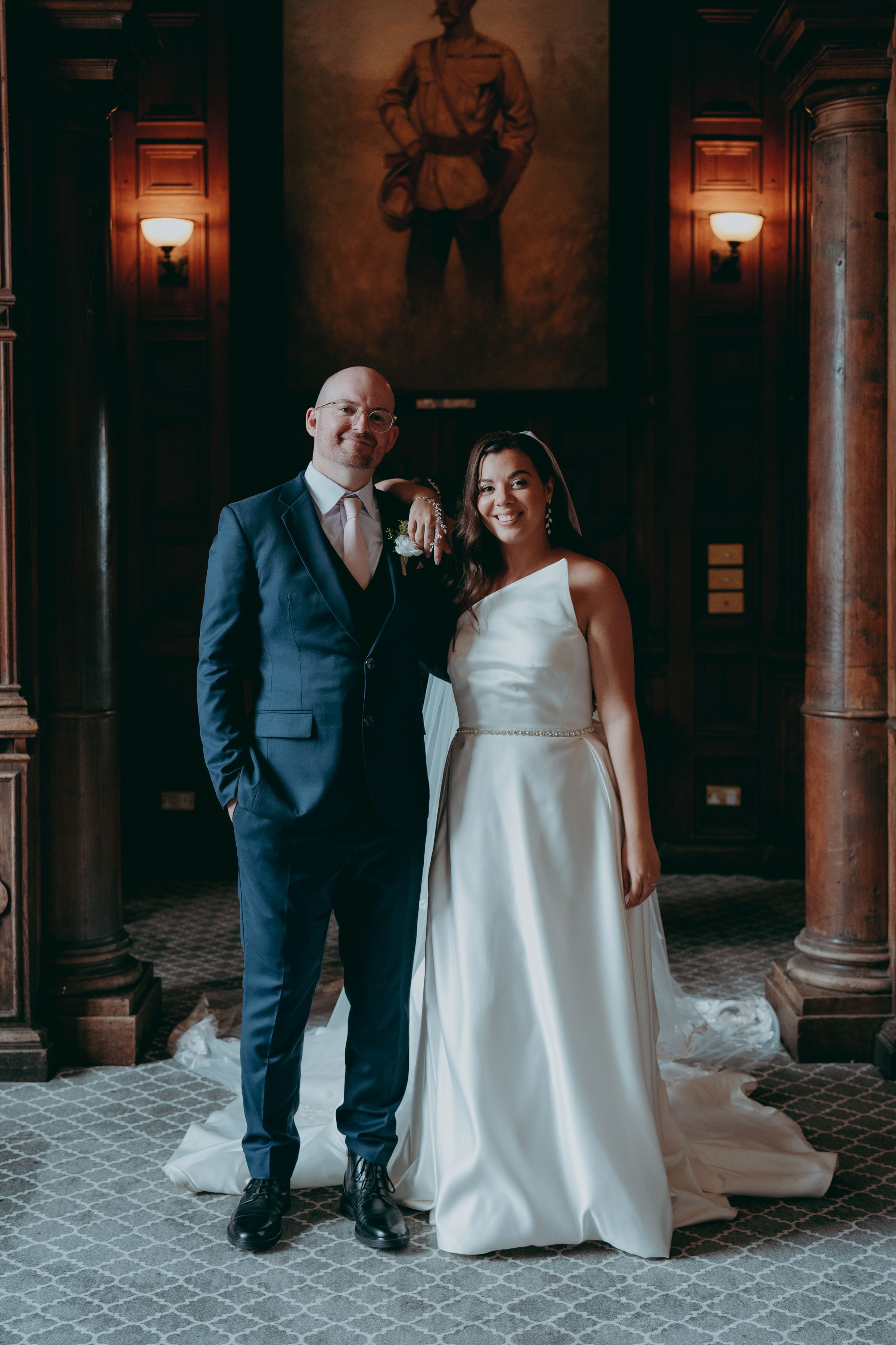 A couple poses for a wedding photo in an elegant venue with wood columns and wall sconces beneath a large painting.