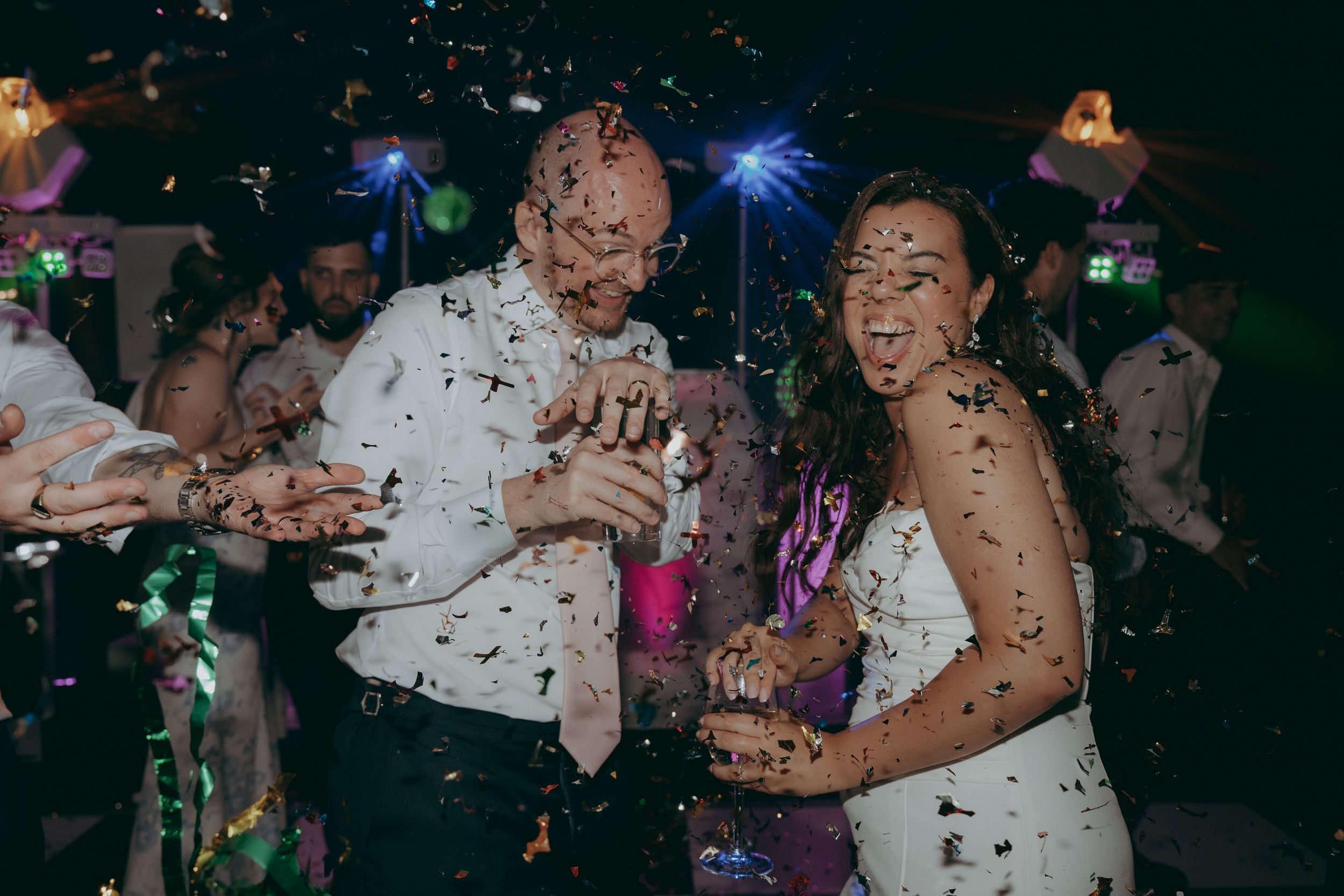 Wedding reception dance party with colorful lights and people celebrating on a dark dance floor with confetti.