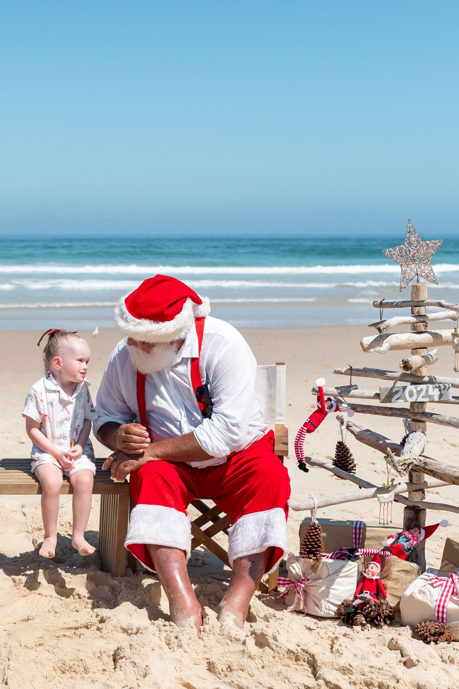 A beach scene at Christmas with Santa sitting on sand next to a driftwood tree decorated with festive ornaments.