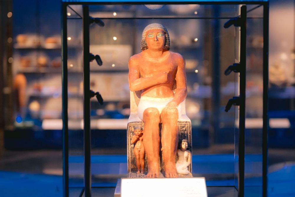 Ancient Egyptian limestone statue on display in a museum case with dramatic lighting.