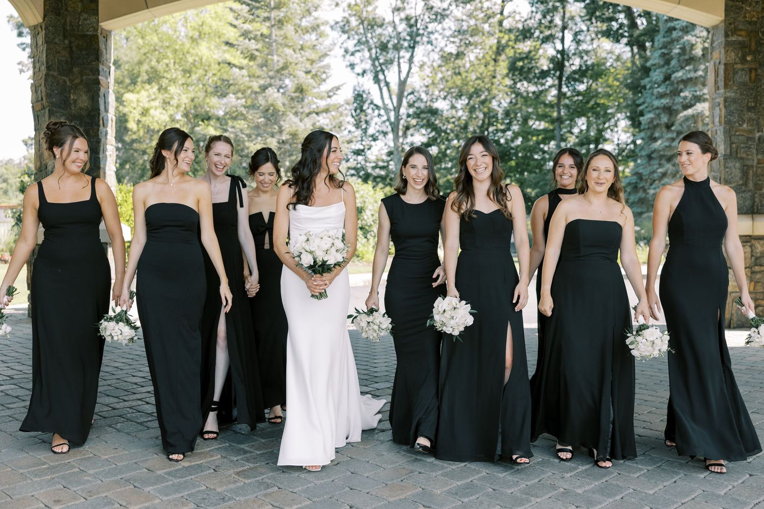 Bridal party stands outdoors wearing elegant black floor-length gowns and one white wedding dress holding white bouquets.