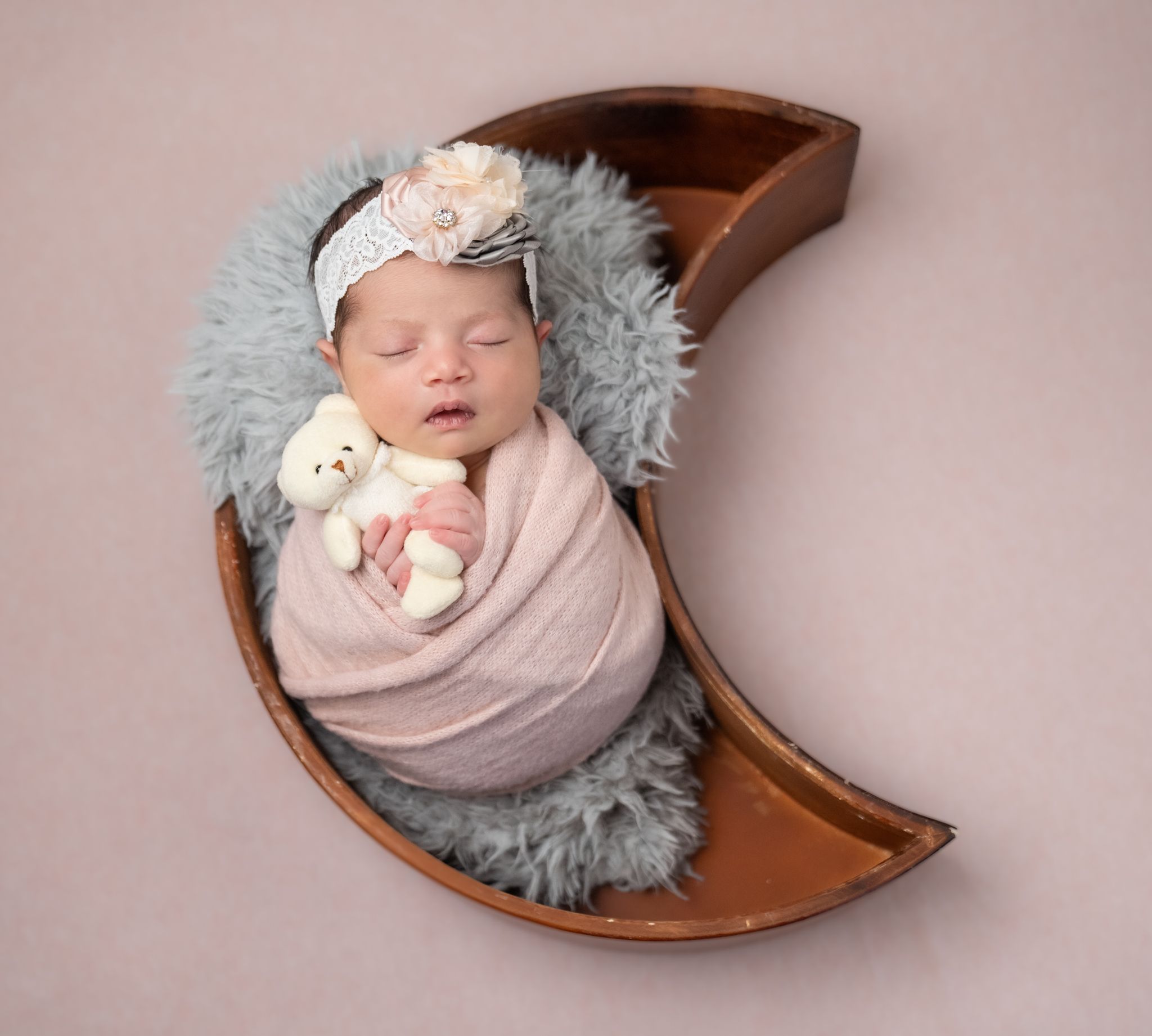 Sleeping newborn swaddled in pink wrap with plush toy rests in wooden crescent moon prop with gray fur padding.