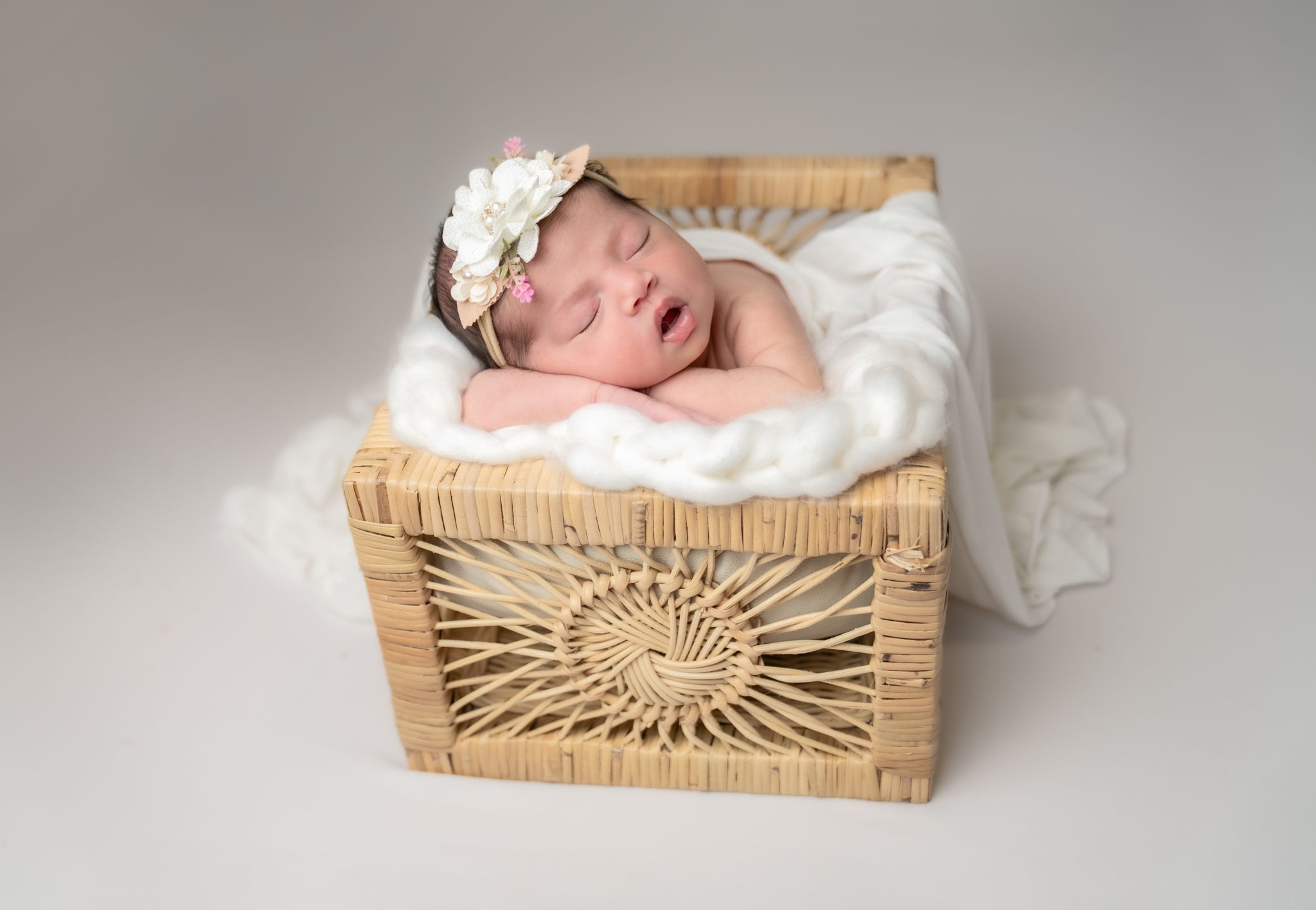 A sleeping newborn rests in a woven basket on soft white fabric, wearing a delicate white floral headband.