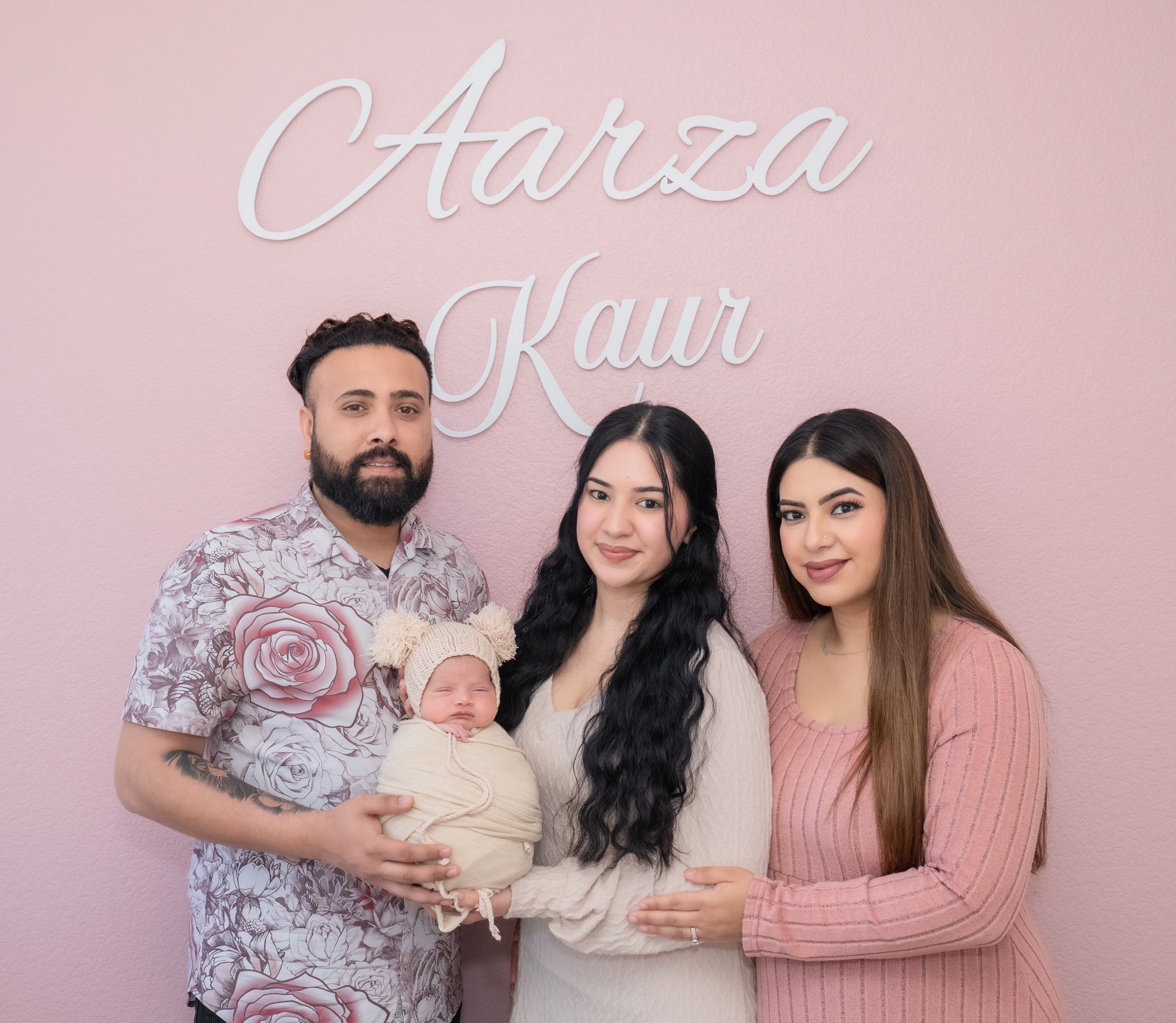 Family portrait against pink background with cursive text, featuring parents holding newborn baby in white outfit.