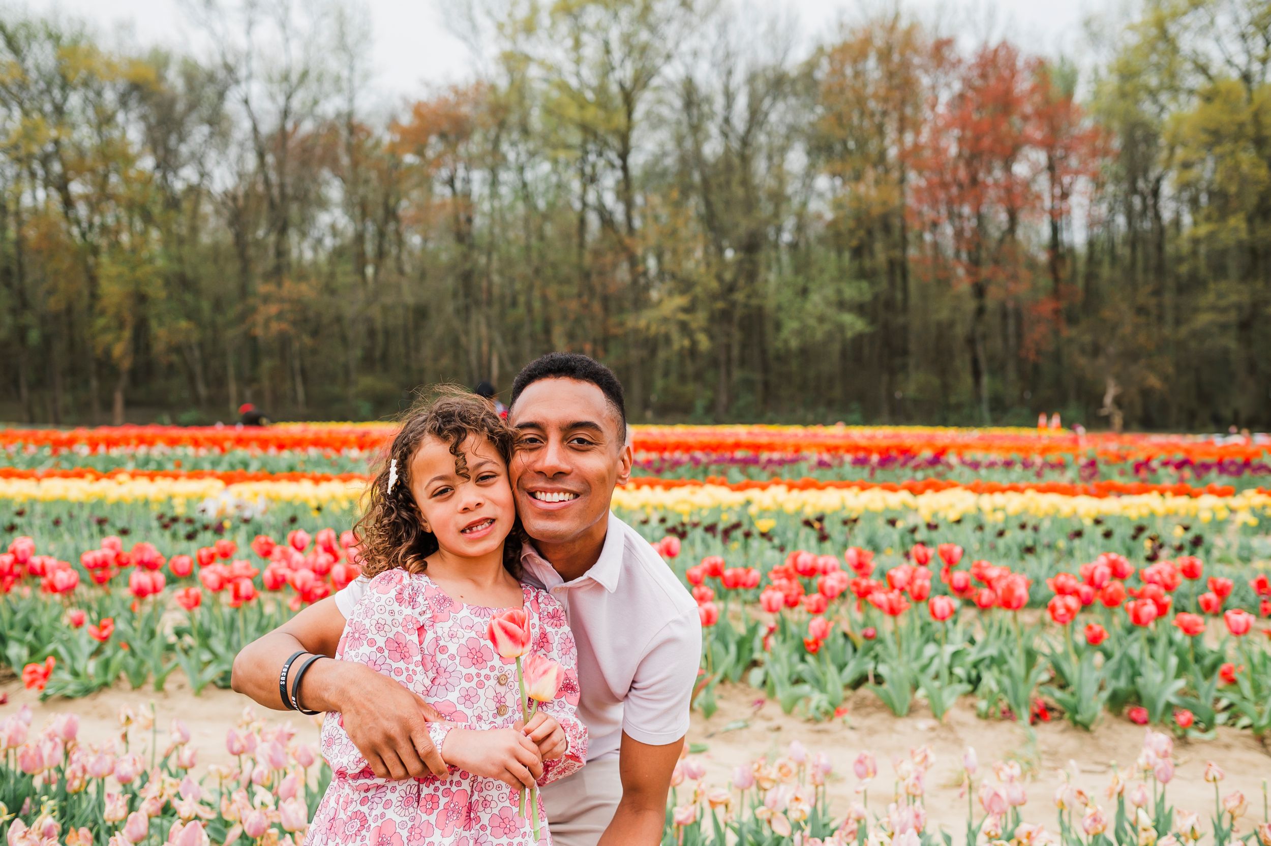 A couple poses together in a colorful tulip field with autumn trees in the background during springtime.