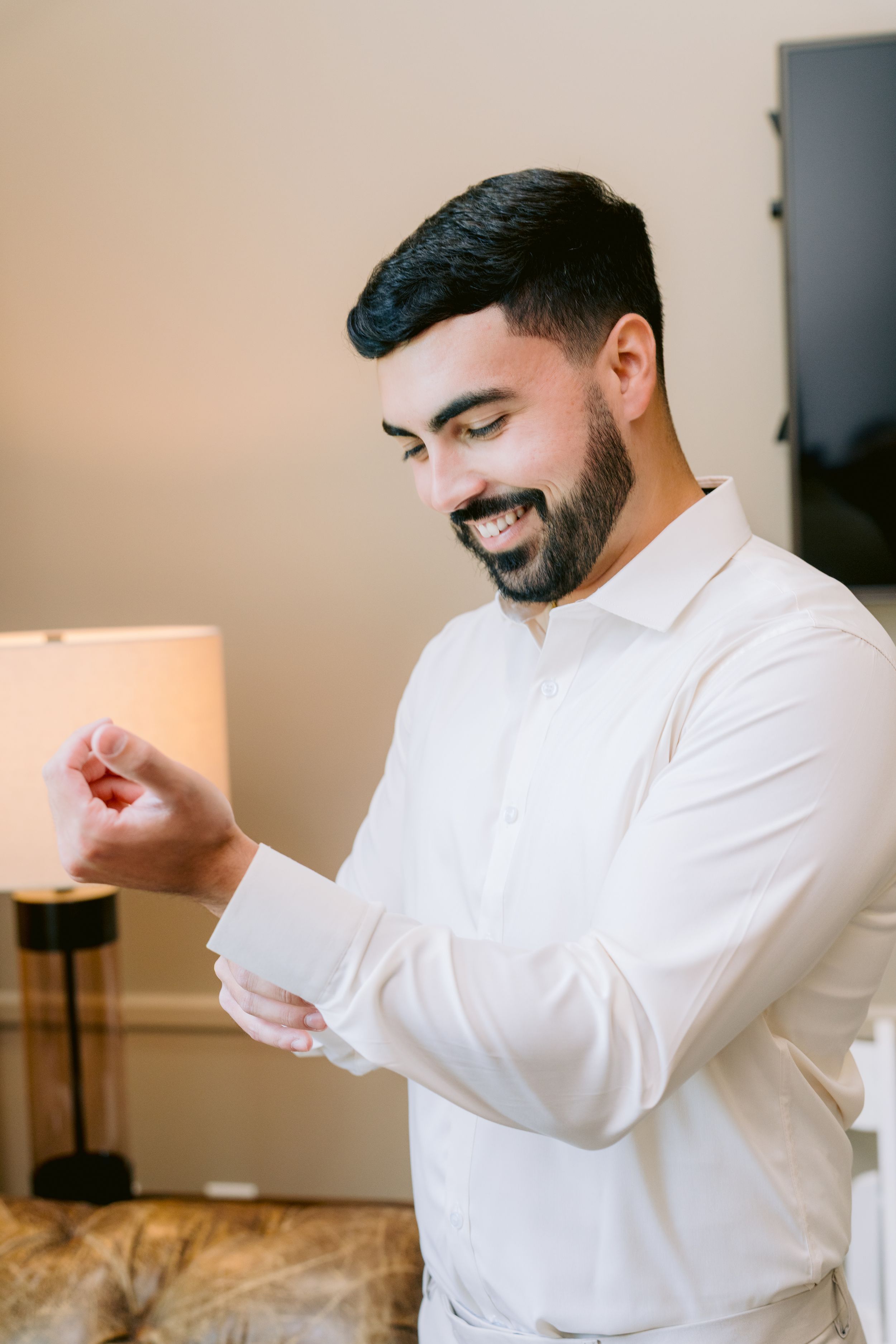 A person in a white dress shirt smiling while adjusting their sleeve cuff in a cozy interior setting.