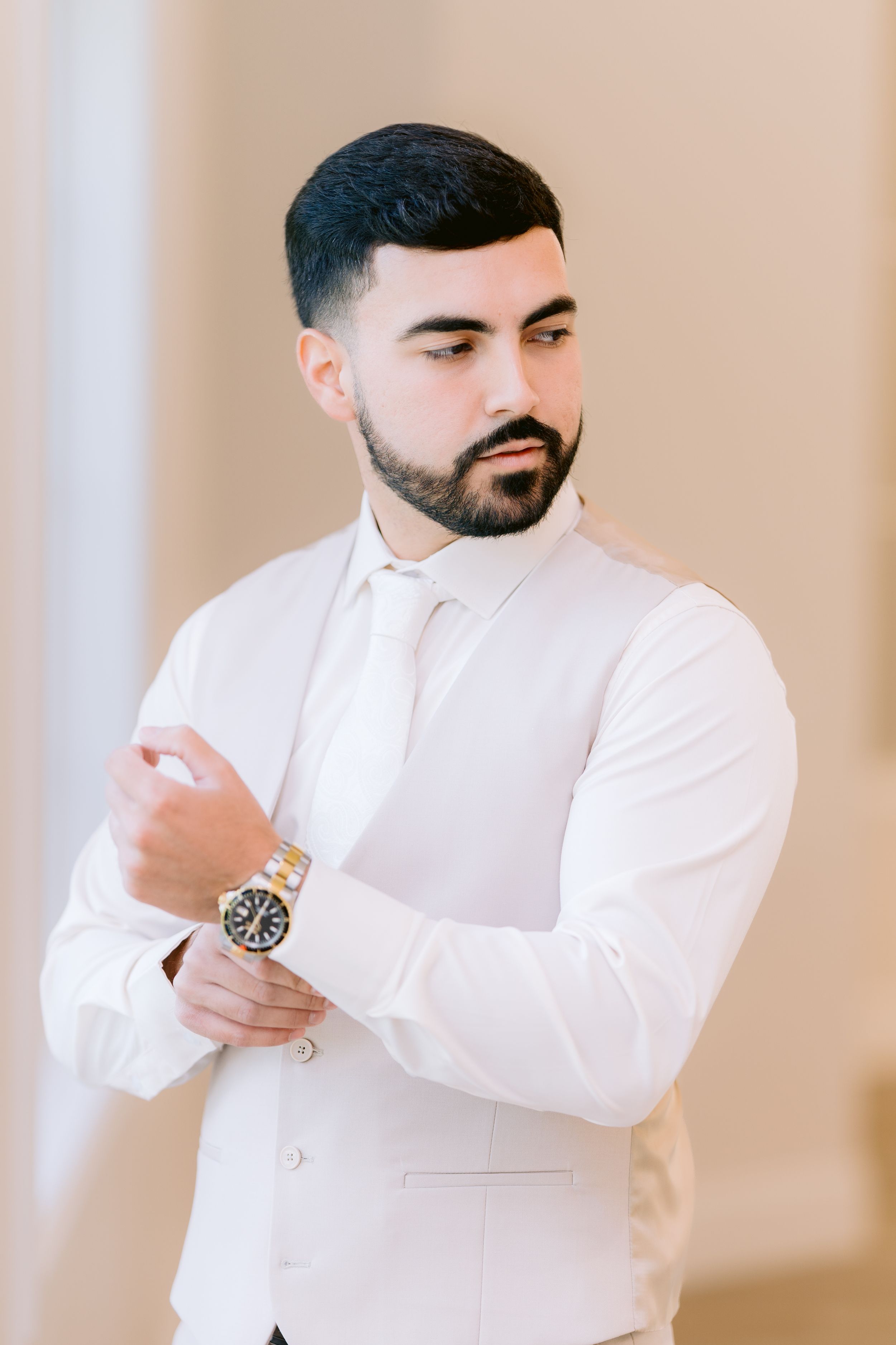Professional in white dress shirt with beard adjusts sleeves while modeling in studio against neutral backdrop.
