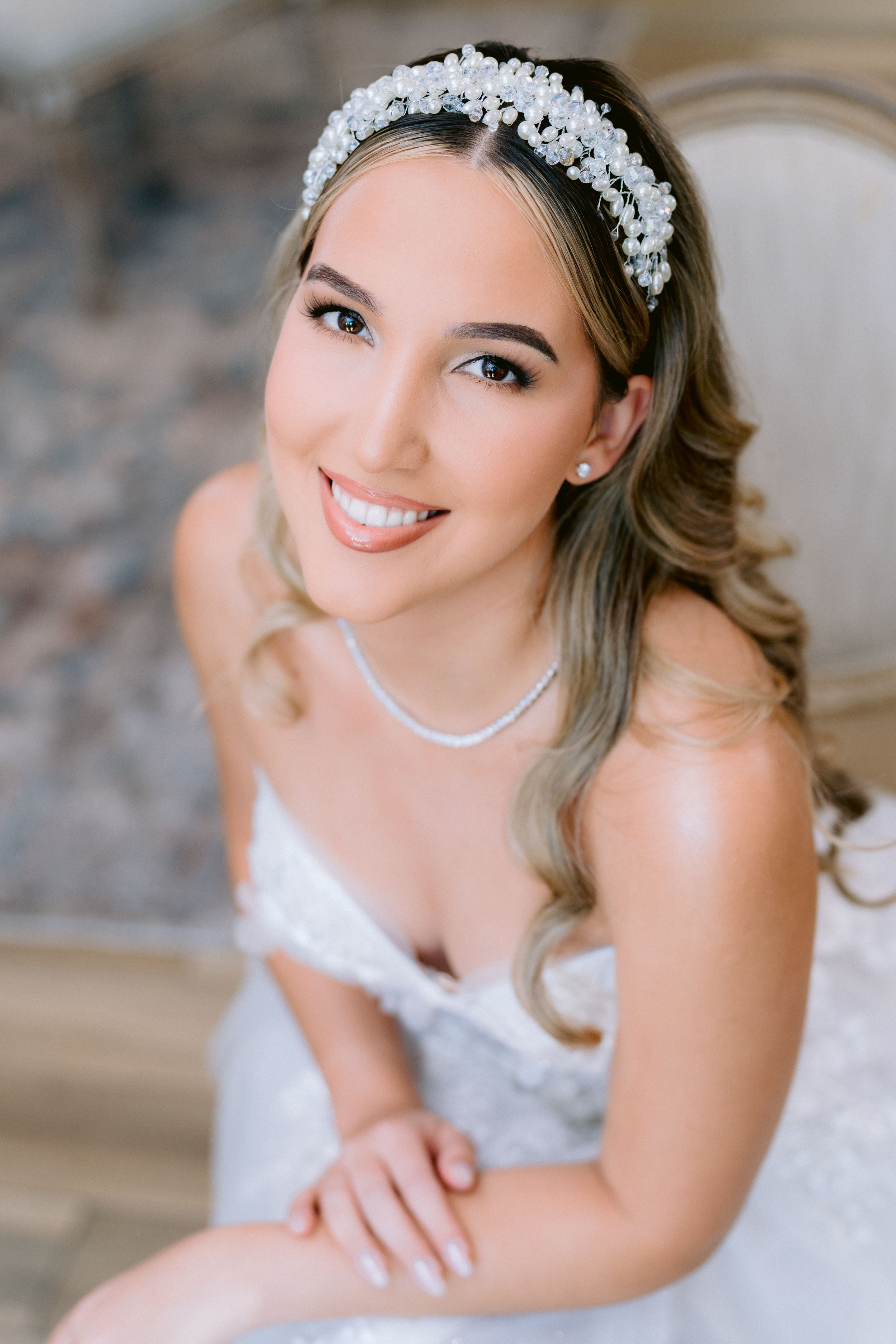 A bride wearing a crystal headpiece and white dress poses for a portrait with soft wavy hair and pearl necklace.