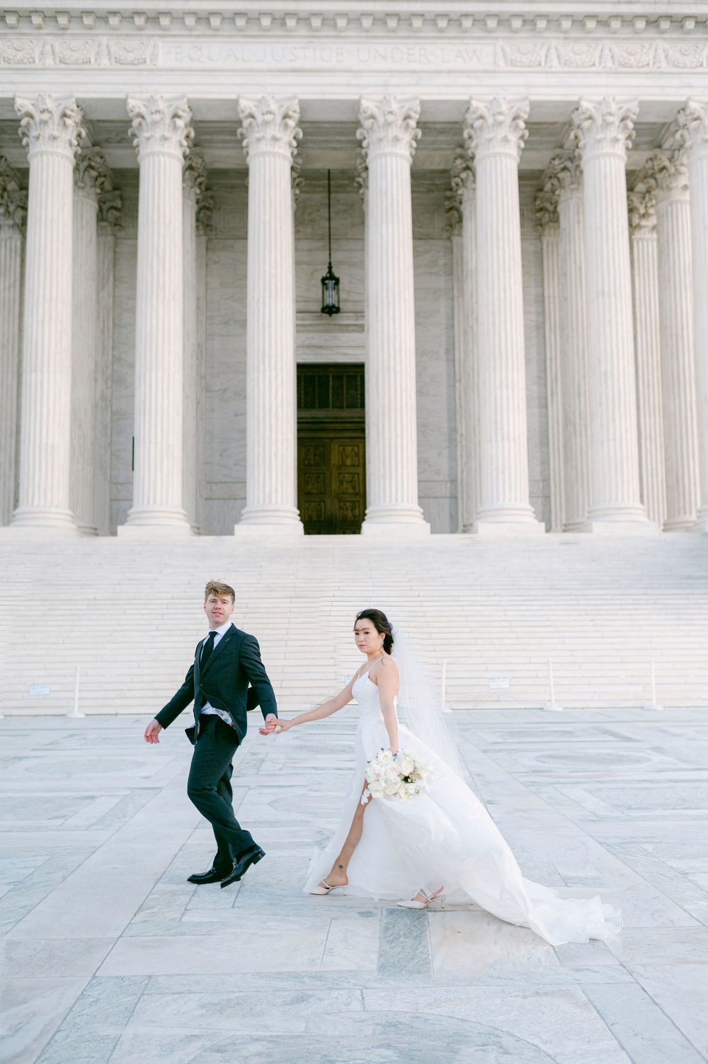 Couple playfully running in front of Supreme Court building with grand white columns.