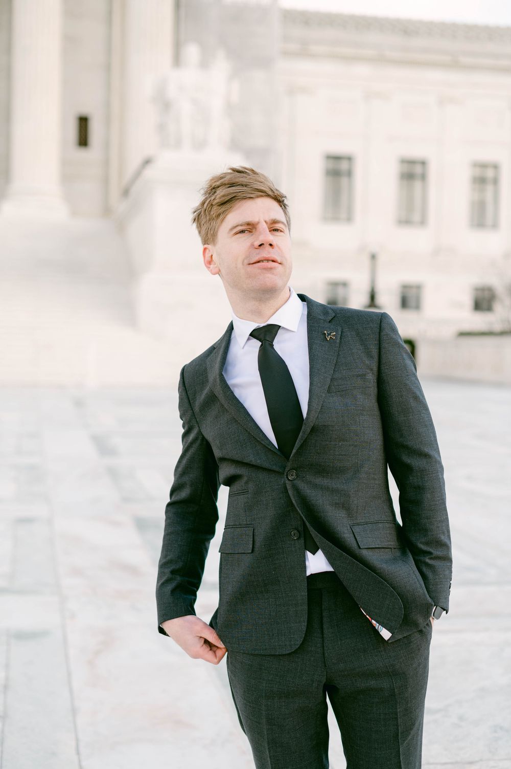 Groom in dark suit and tie posing against white stone building.