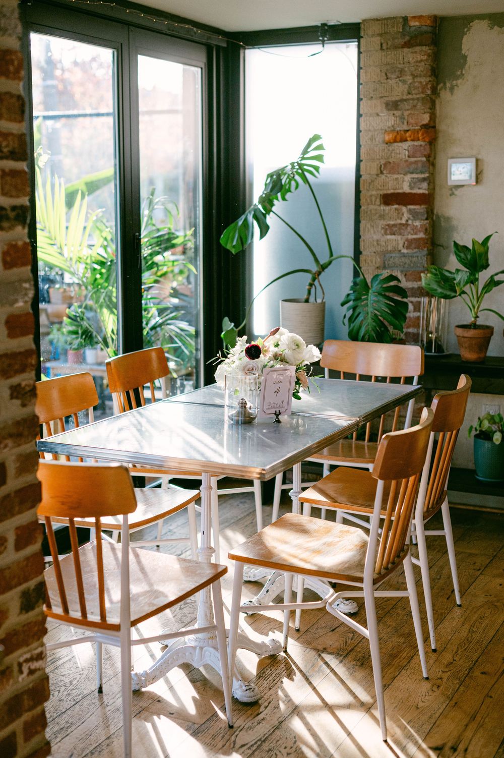 Sunlit dining room with wooden chairs and houseplants around white table.