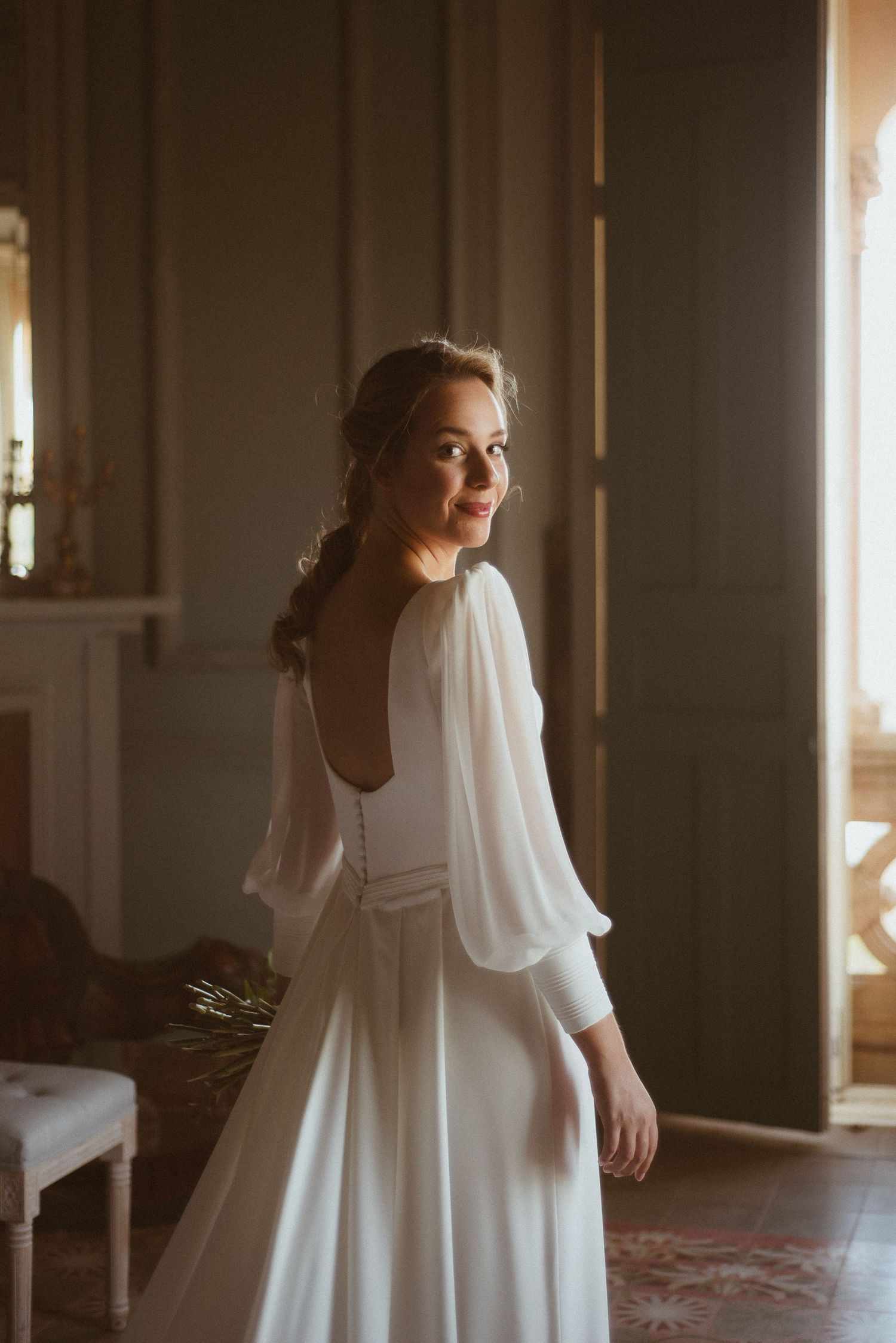 A bride in a flowing white dress with billowing sleeves stands in warm sunlight streaming through a window.