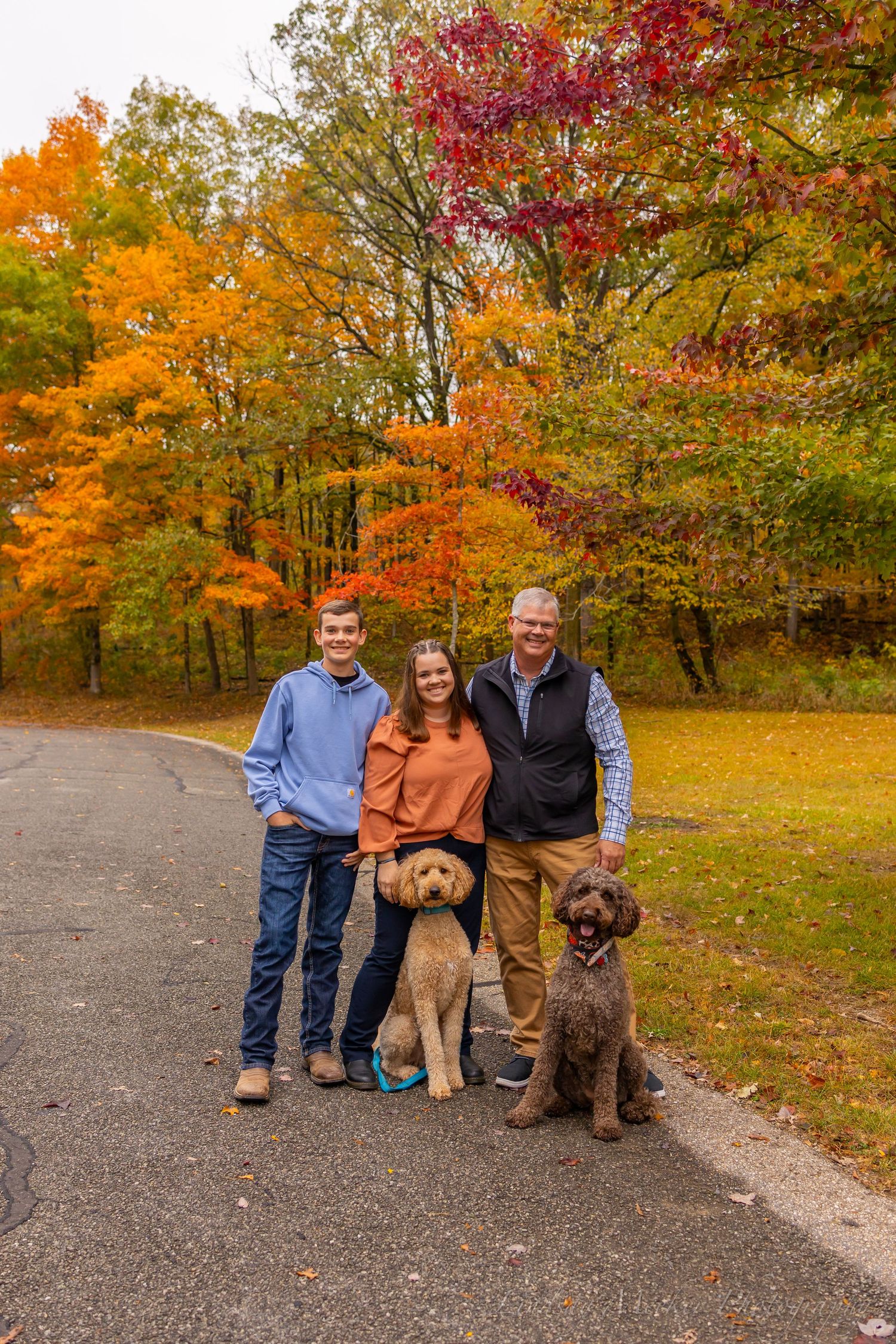 Three people and two dogs pose together on a path surrounded by vibrant fall foliage in orange, red and yellow.