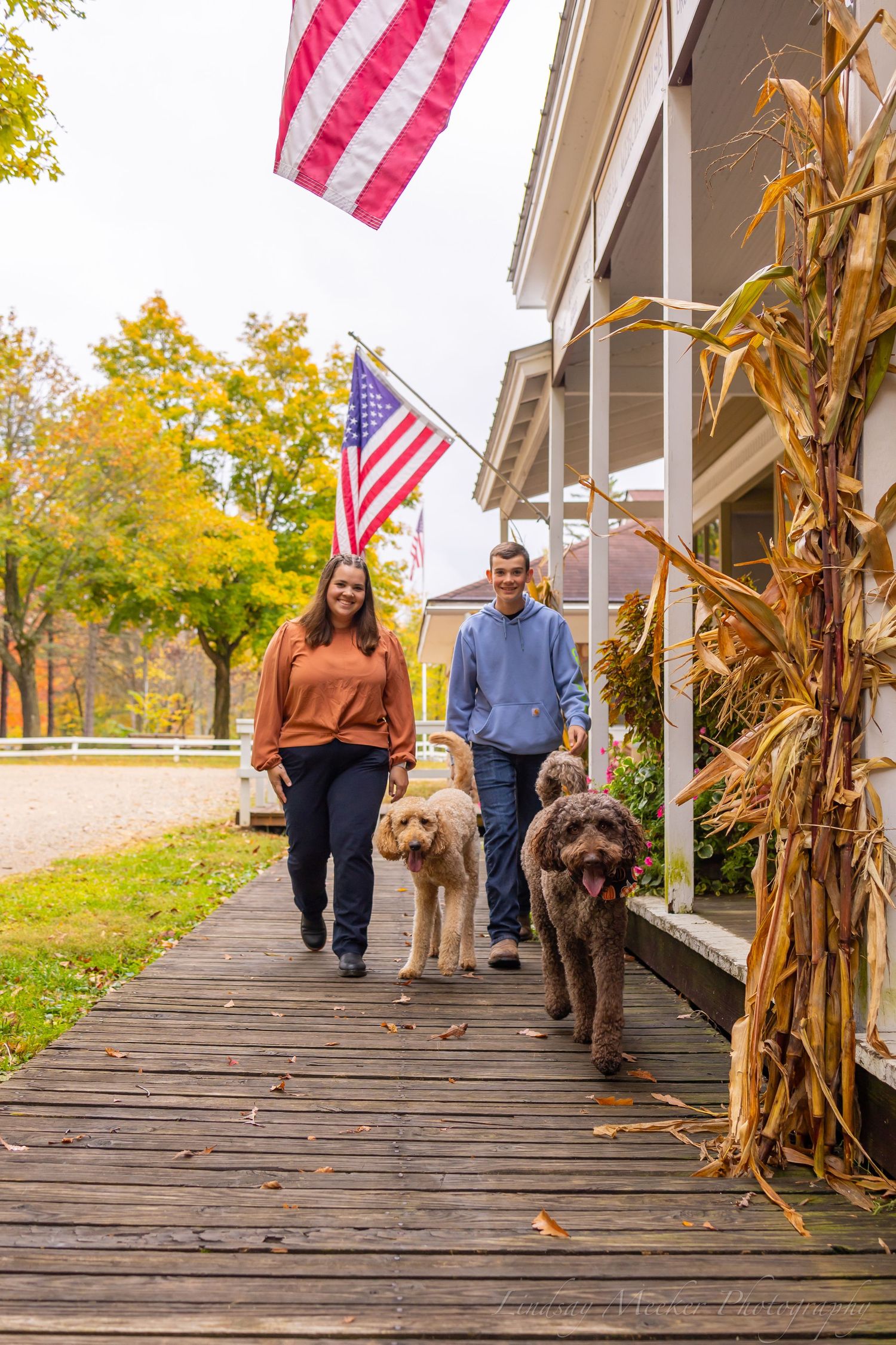 Couple walking a golden retriever on wooden porch decorated with fall cornstalks and American flag.