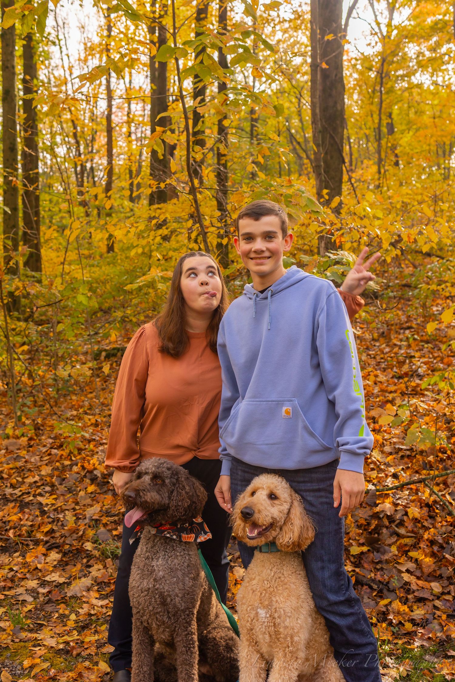 Two people pose with their Airedale terrier dogs among vibrant fall foliage in a forest setting.