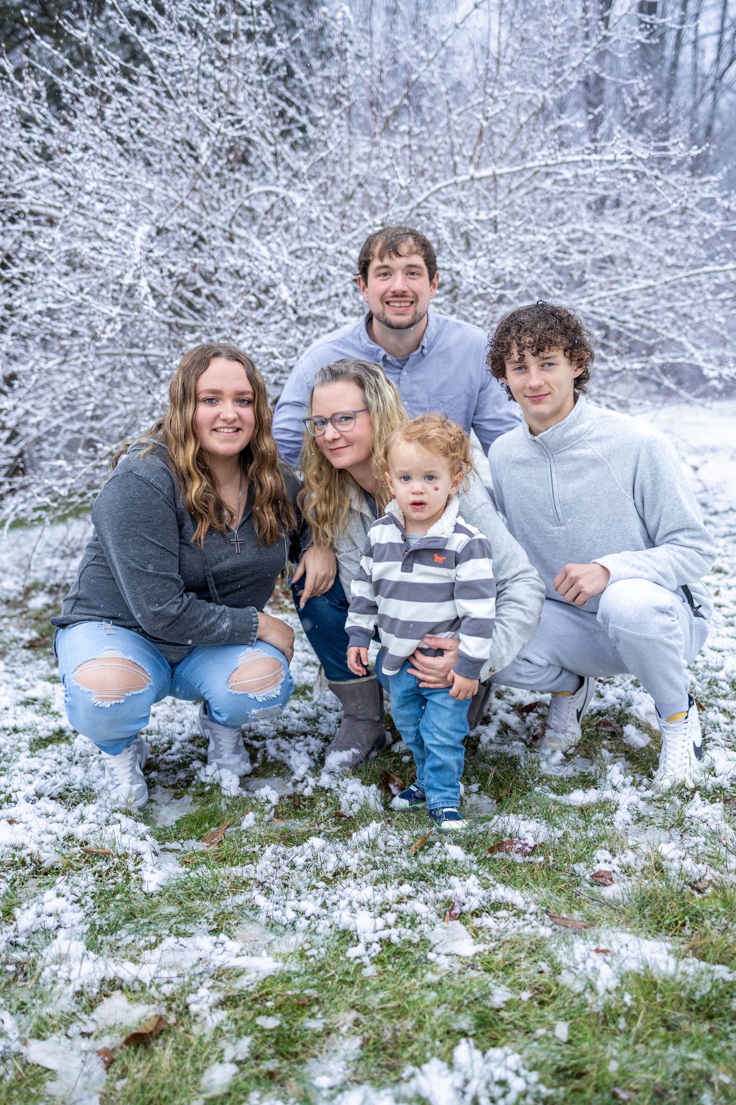 Group sits together in snowy forest setting, smiling and posing playfully on white winter ground.