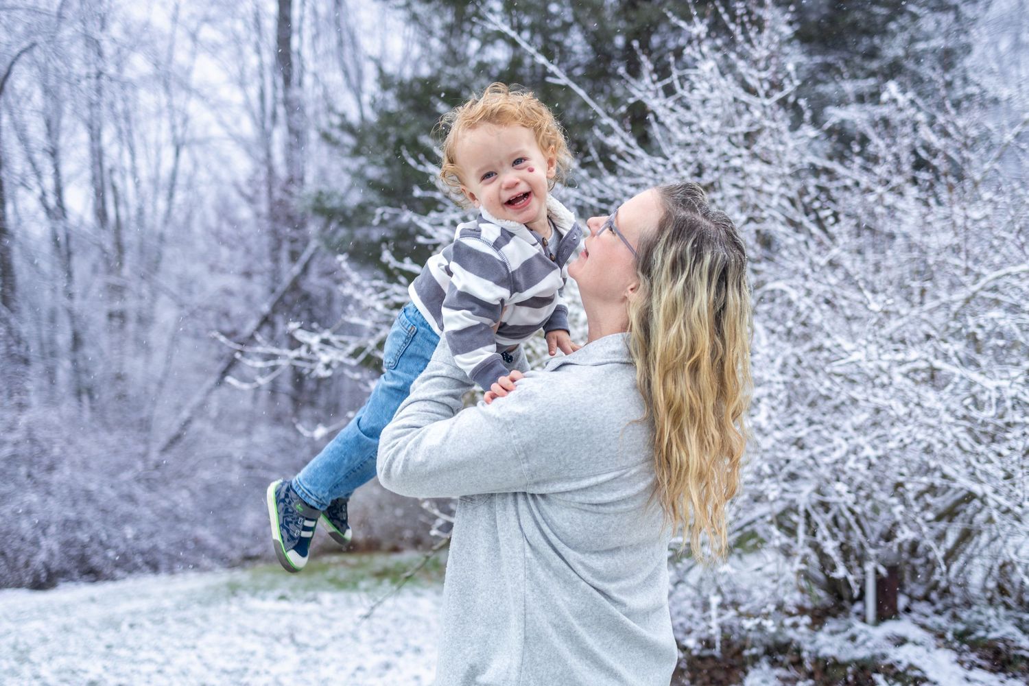 In a snowy winter forest, a parent in teal jacket lifts up a laughing child during a playful family photo session.