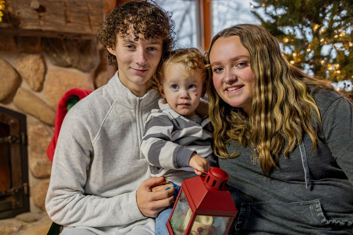 Three people sit together by a stone wall while holding a red gift box during winter celebrations.