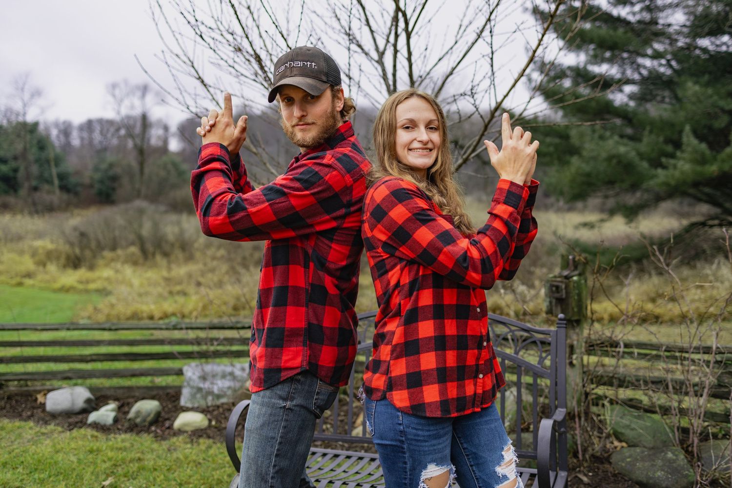 Two people in matching red buffalo plaid flannel shirts pose back-to-back outdoors by a wooden fence.