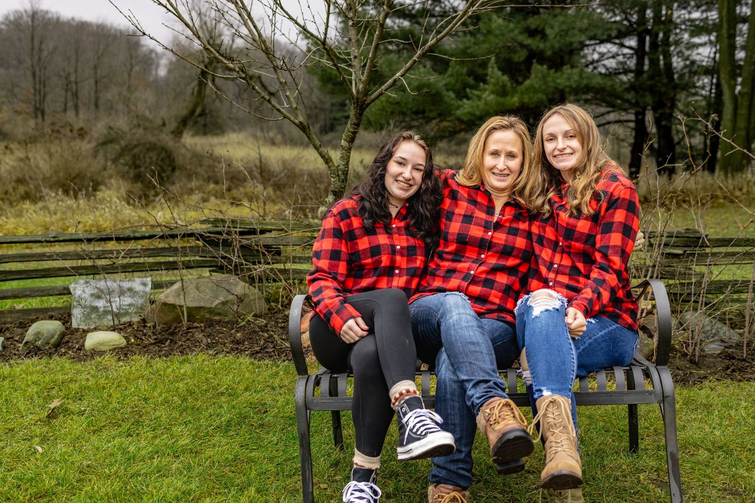 Three people wearing matching red and black buffalo plaid shirts sit together on an outdoor bench by a pond.