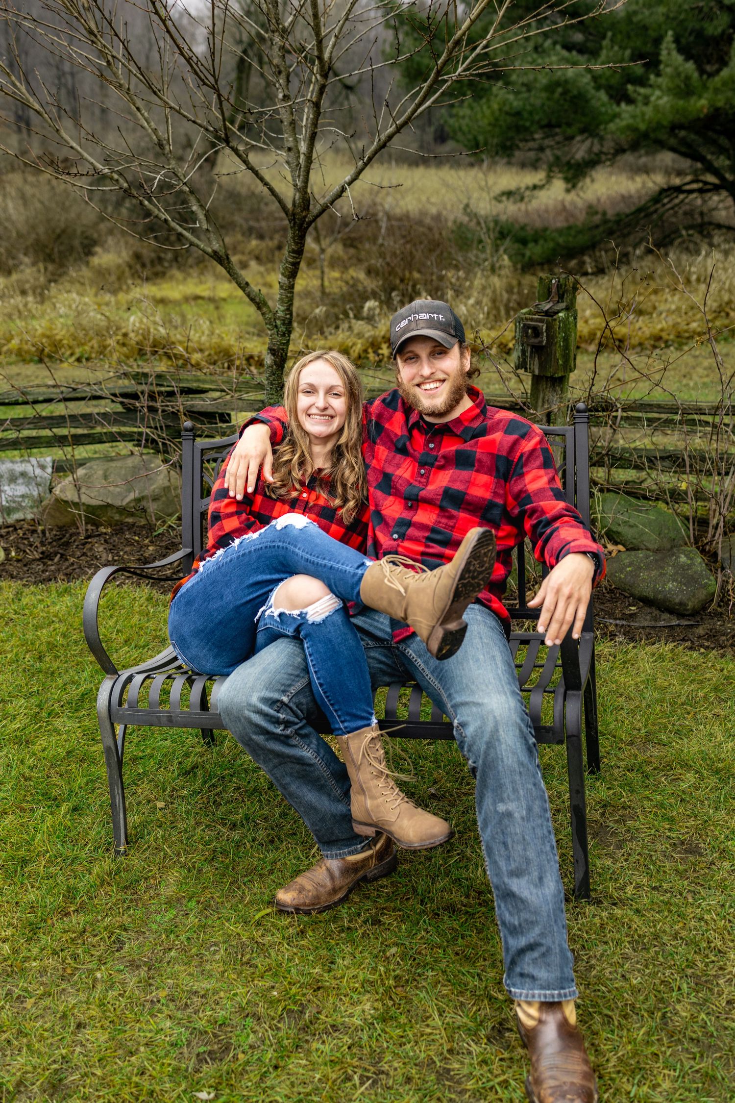 A couple in casual attire lounging together on a park bench surrounded by green grass and trees.
