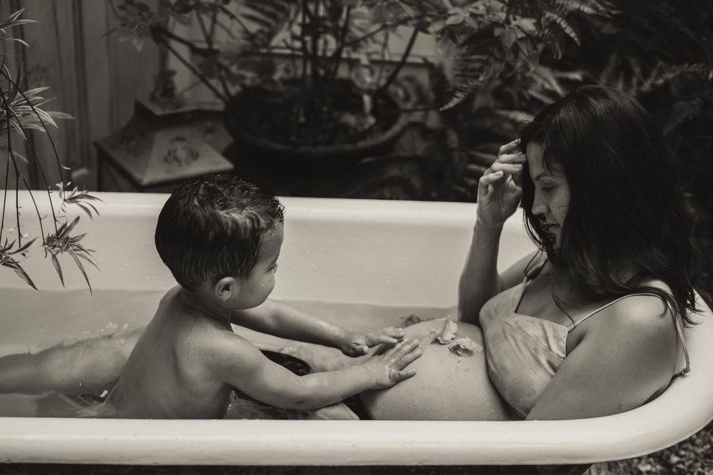 son reaches out to touch his mother's pregnant belly as they sit in a backyard bathtub together after photo session