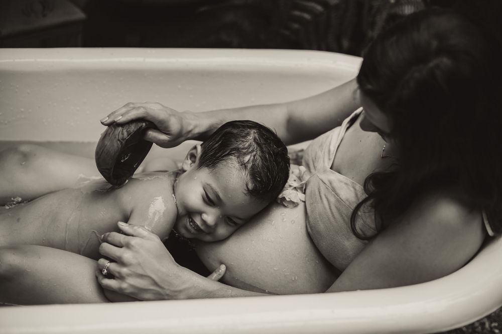 son hugs mother's pregnant belly in a backyard bathtub as she pours water over his back from wooden bowl