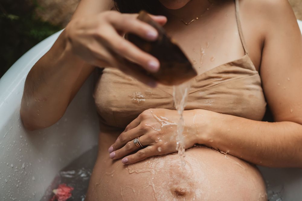 close up of woman in linen top pours water from wooden bowl onto her bare pregnant belly in backyard tub