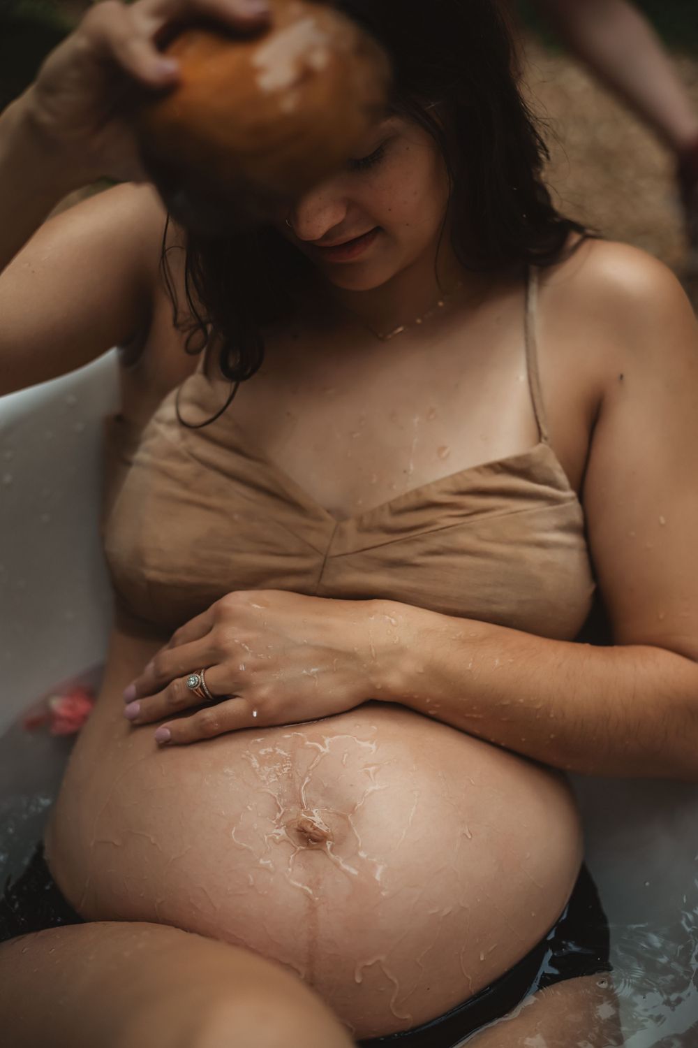woman drips water from wooden bowl onto her pregnant belly in an outdoor bathtub photoshoot