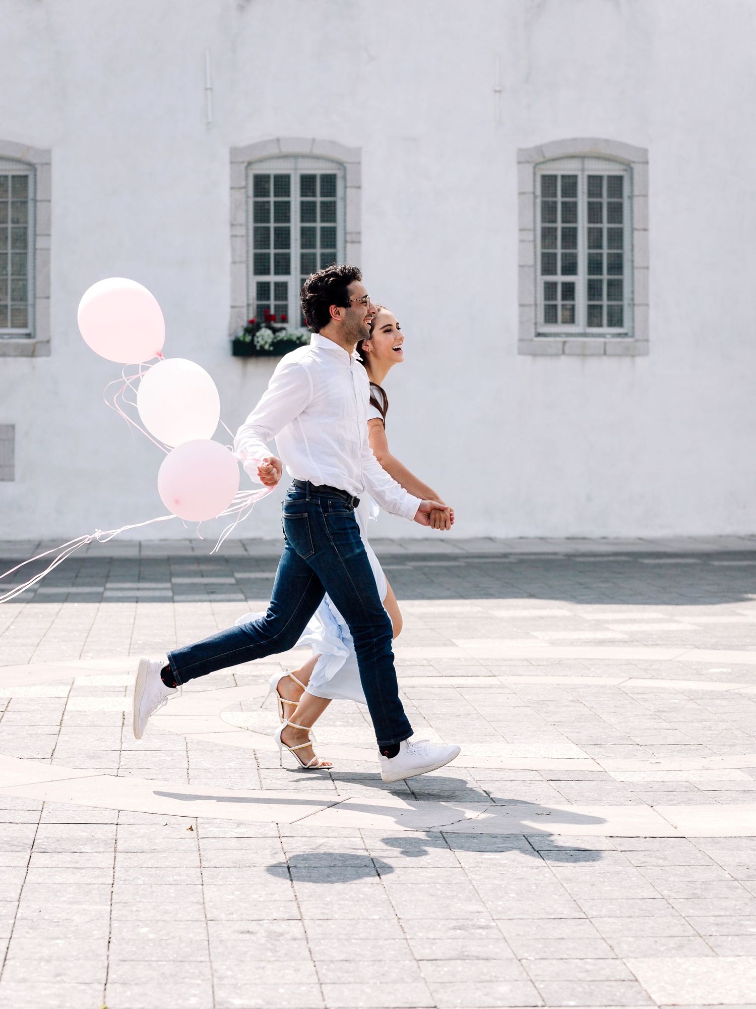 Couple running joyfully with pink balloons against a white building facade.