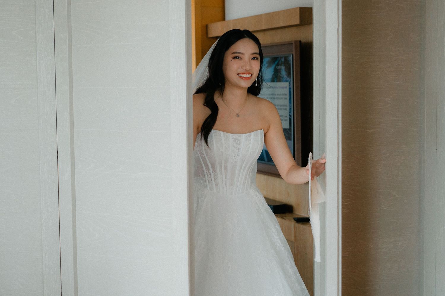 Elegant bride in an off-shoulder white gown stands in a bright doorway with a joyful expression.
