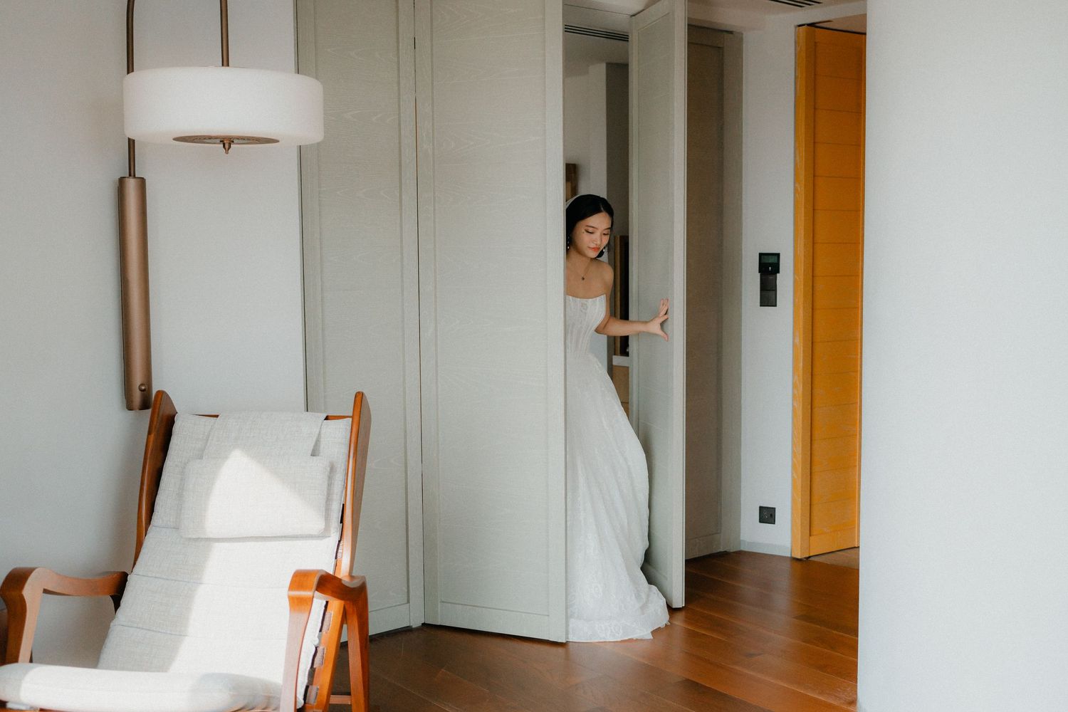 A white bridal gown peeks out from behind a doorway in a modern room with wooden flooring and a white chair.