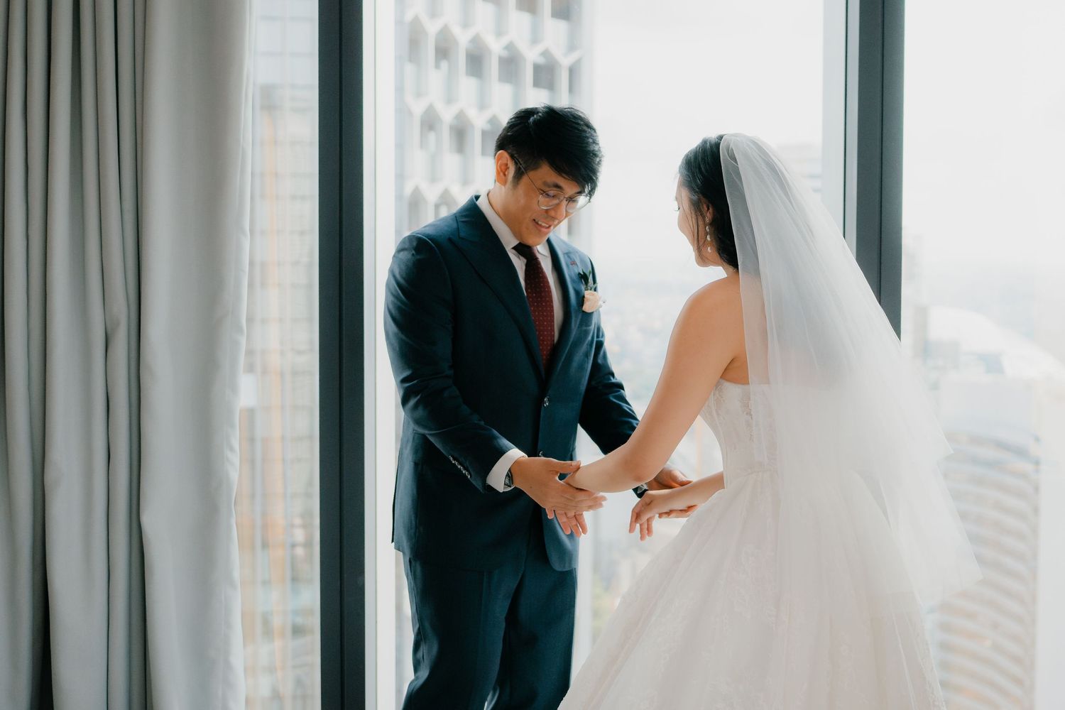 A couple in wedding attire share an intimate moment by a window overlooking a city skyline.
