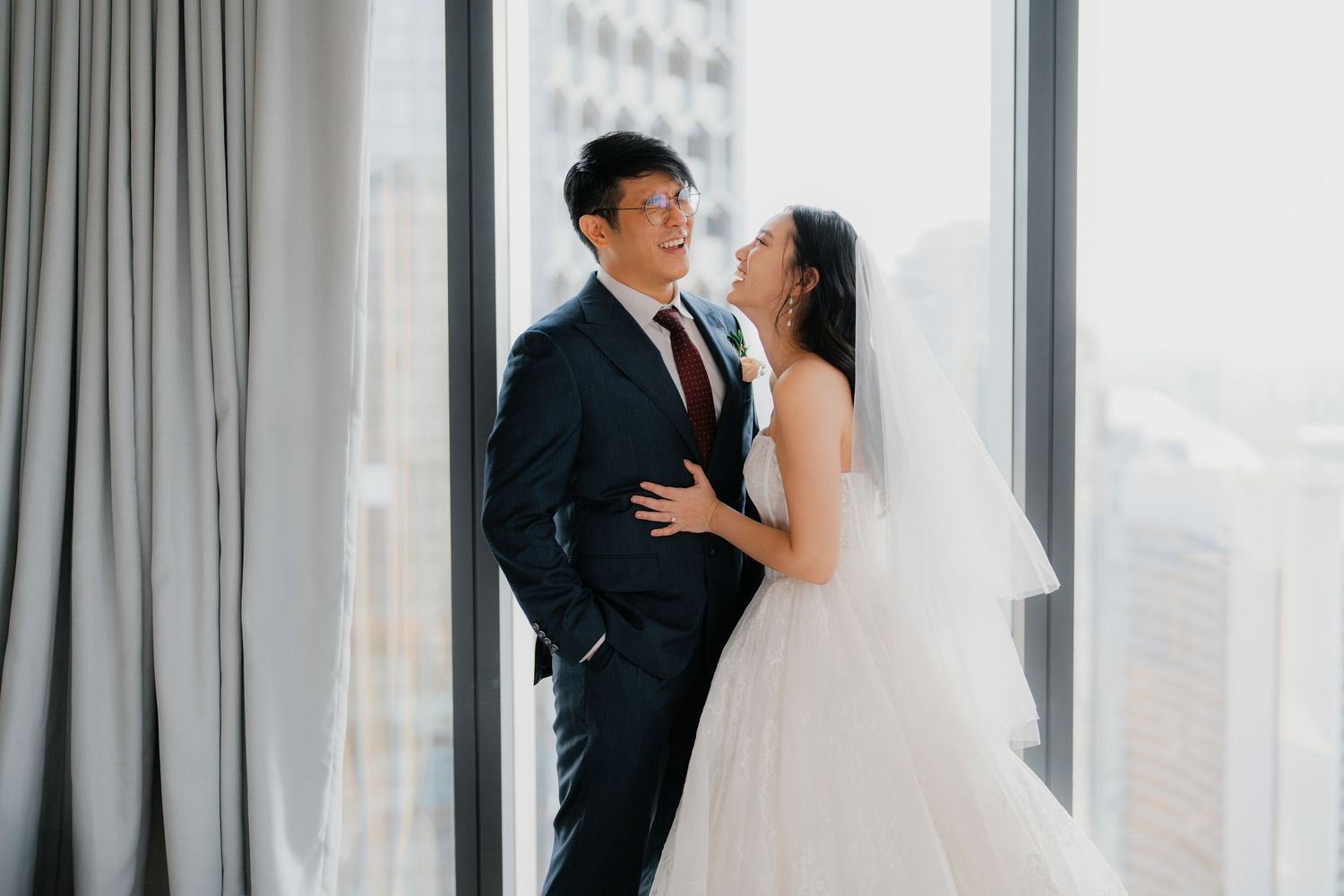 A couple in wedding attire shares an intimate moment by large windows overlooking city buildings.