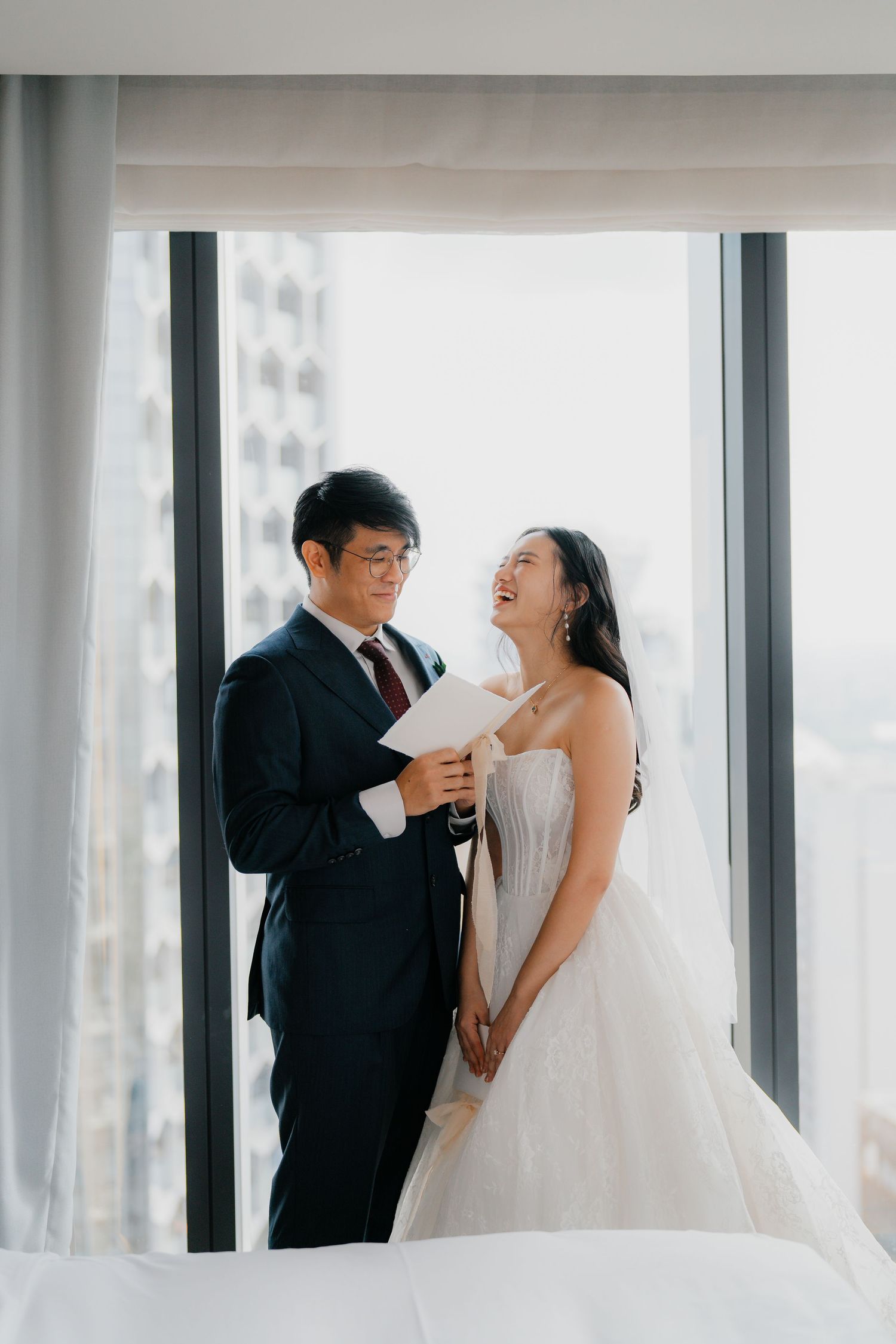 A joyful couple shares a candid moment together by large windows in wedding attire.