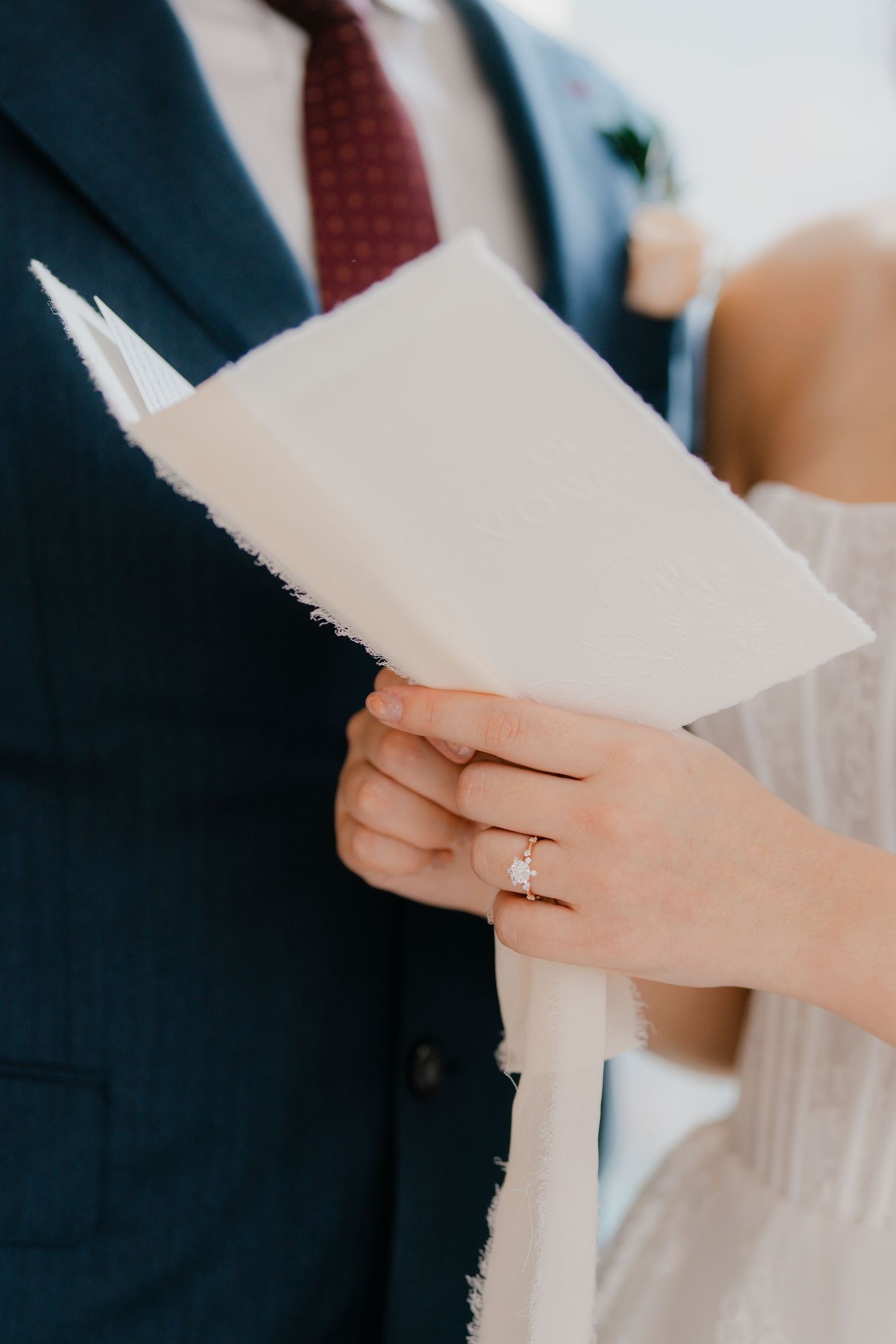 Two hands holding pages of a wedding ceremony script, with a diamond engagement ring visible on the bride's finger.