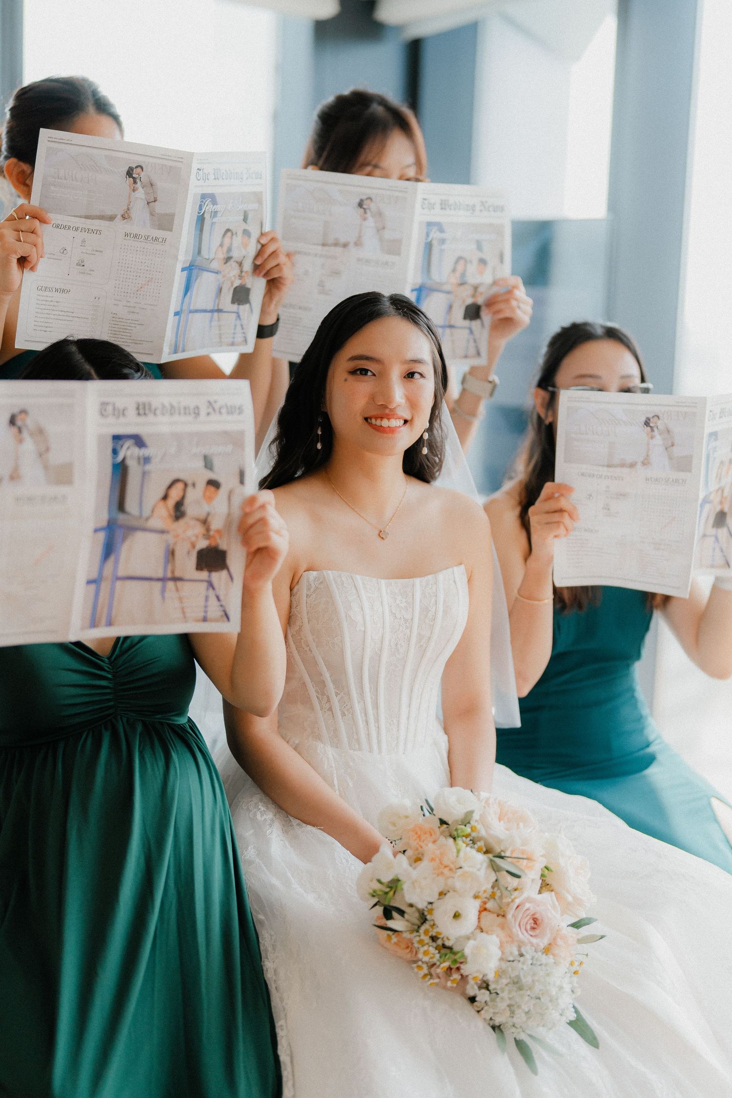 A bride in white dress poses with bridesmaids in emerald green dresses holding up papers during wedding preparation.