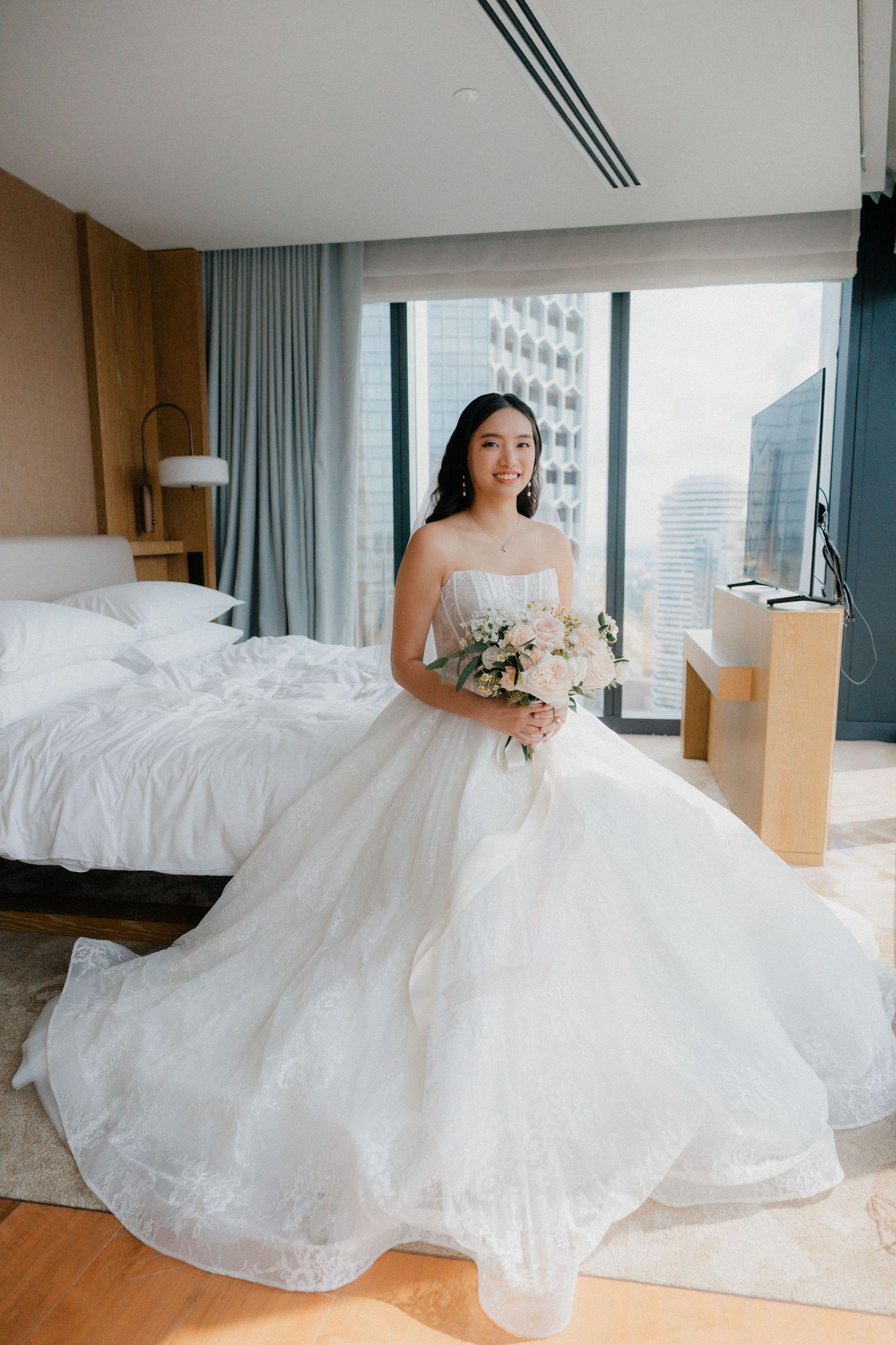 A bride in a flowing white ballgown poses in a luxurious hotel room with floor-to-ceiling windows and city views.