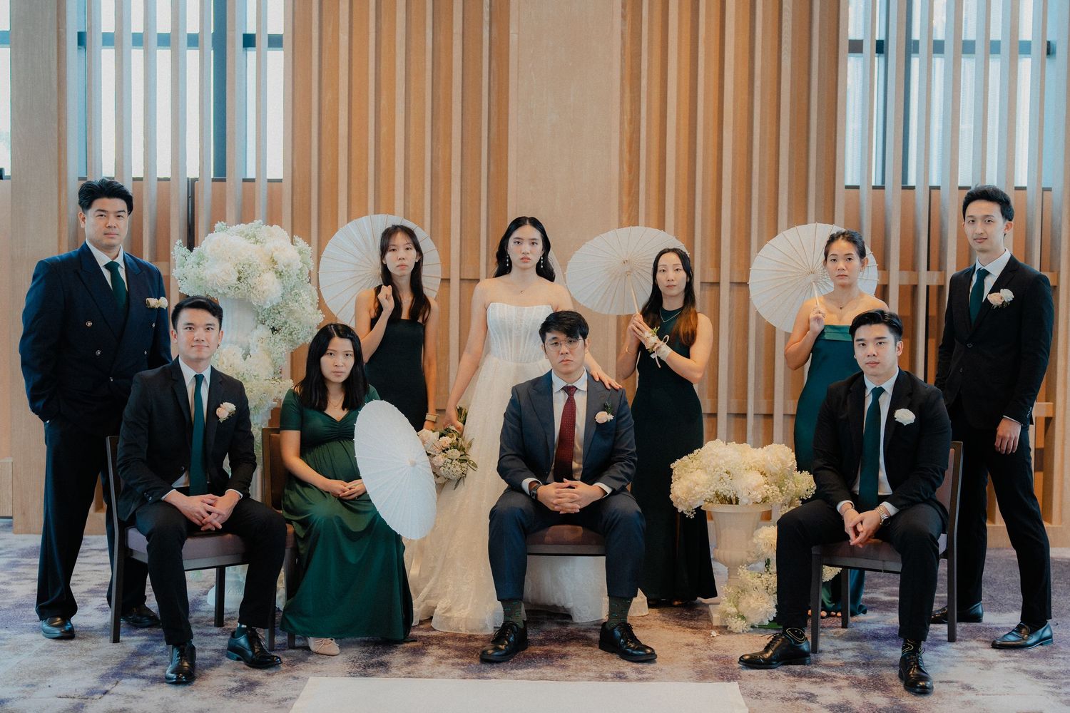 Wedding party members pose formally in emerald green dresses and dark suits against a wooden panel wall.