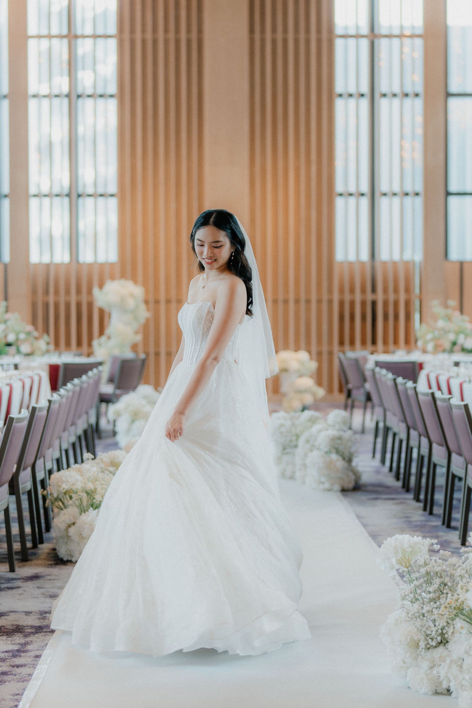Elegant bride in white gown posing gracefully in a wedding venue decorated with delicate white floral arrangements.