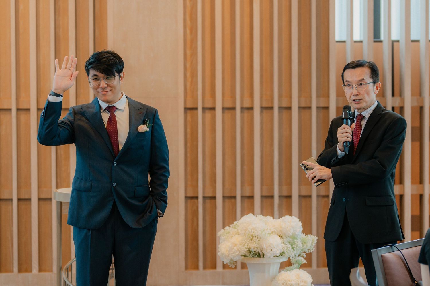 Two speakers in formal attire present during an indoor wedding ceremony.