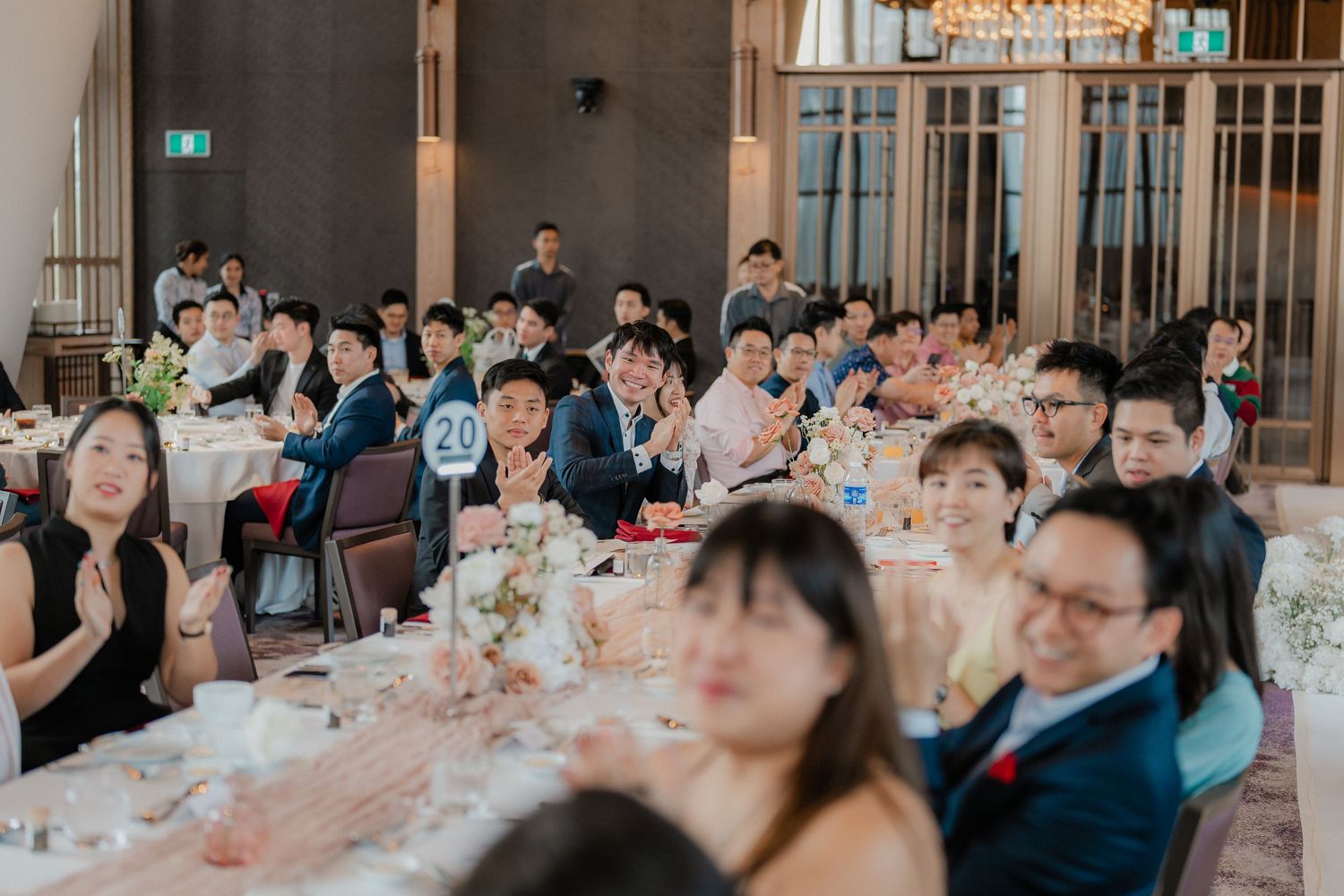 Wedding guests sit at elegant tables decorated with floral centerpieces during an indoor reception celebration.