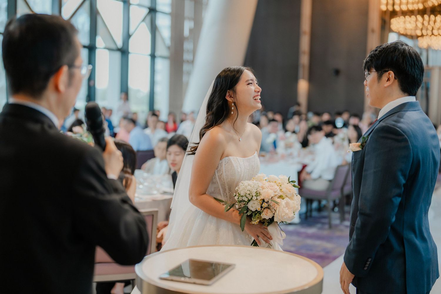 Wedding ceremony moment in a bright venue with chandeliers and large windows.