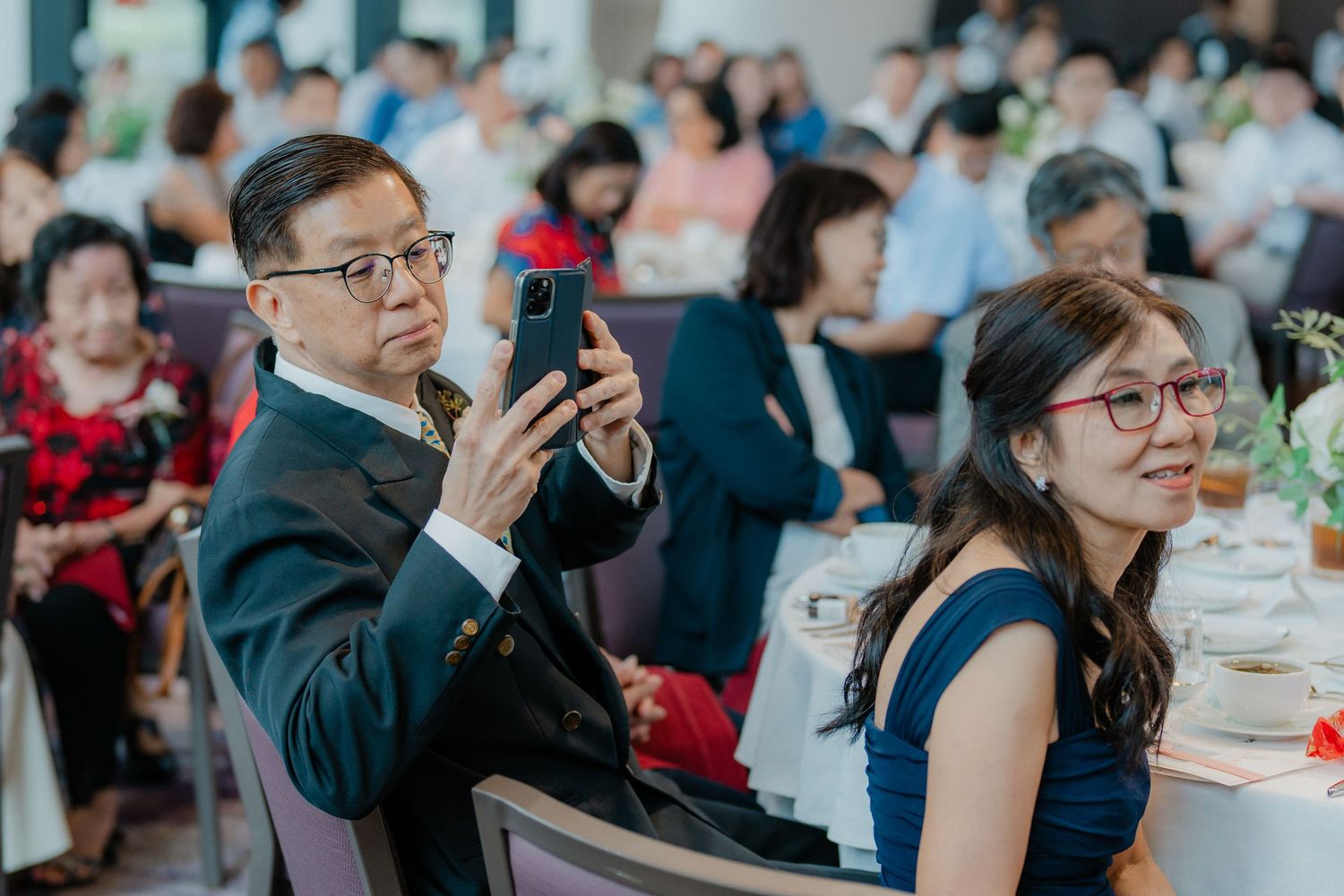 Wedding guests at a banquet table with one person taking photos on a mobile phone.